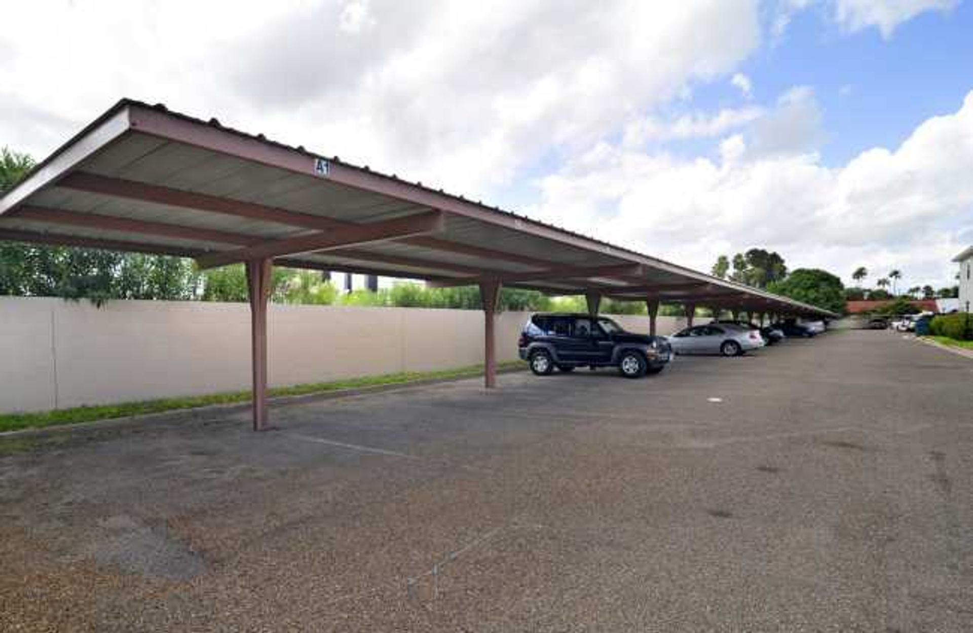 Covered parking lot with several parked cars beside a white wall under a cloudy sky