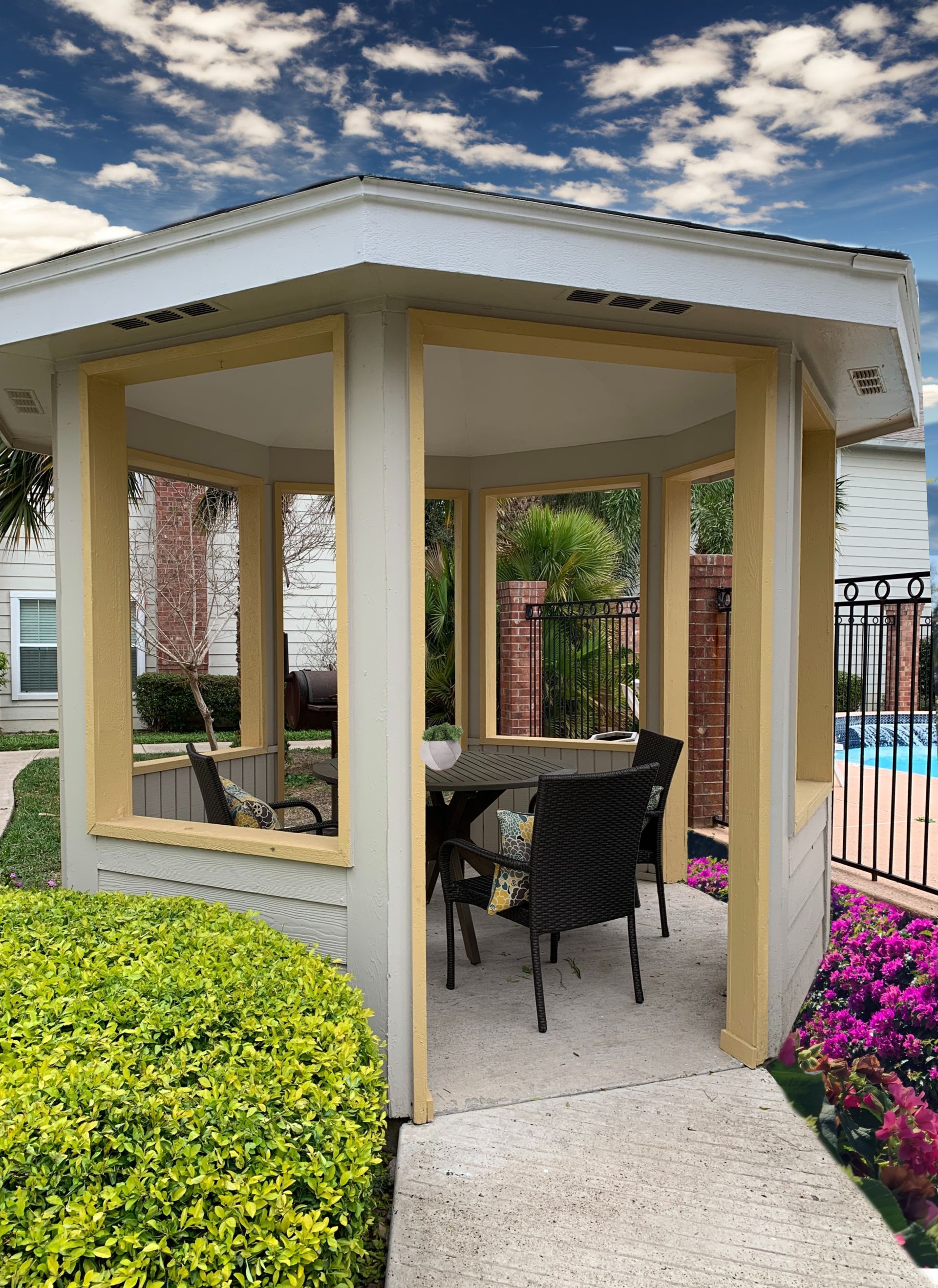 Covered patio gazebo with wicker chairs beside a pool and flowers under a partly cloudy sky