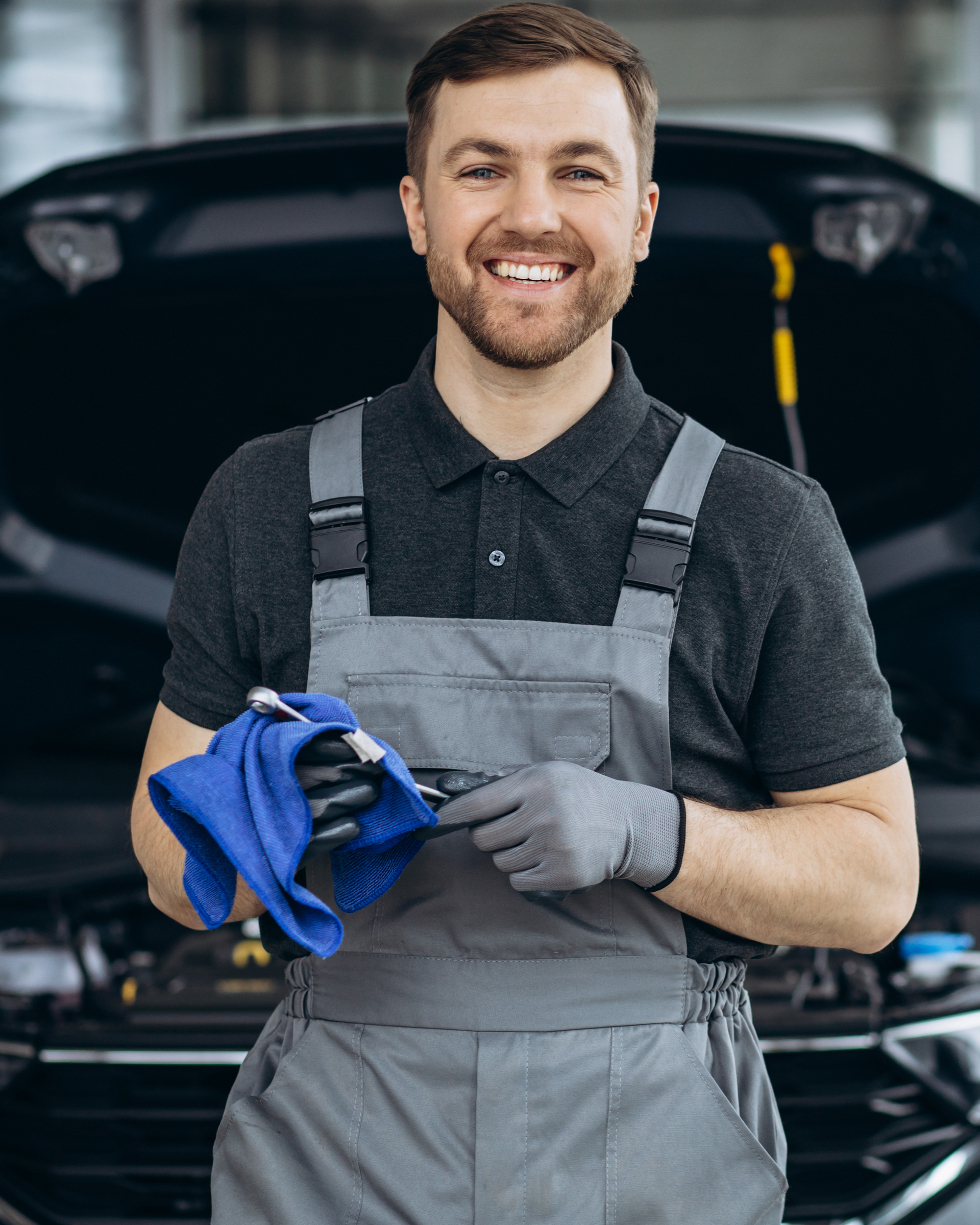 Smiling mechanic holding wrench and rag, standing in front of an open car hood. | Railside Auto Service