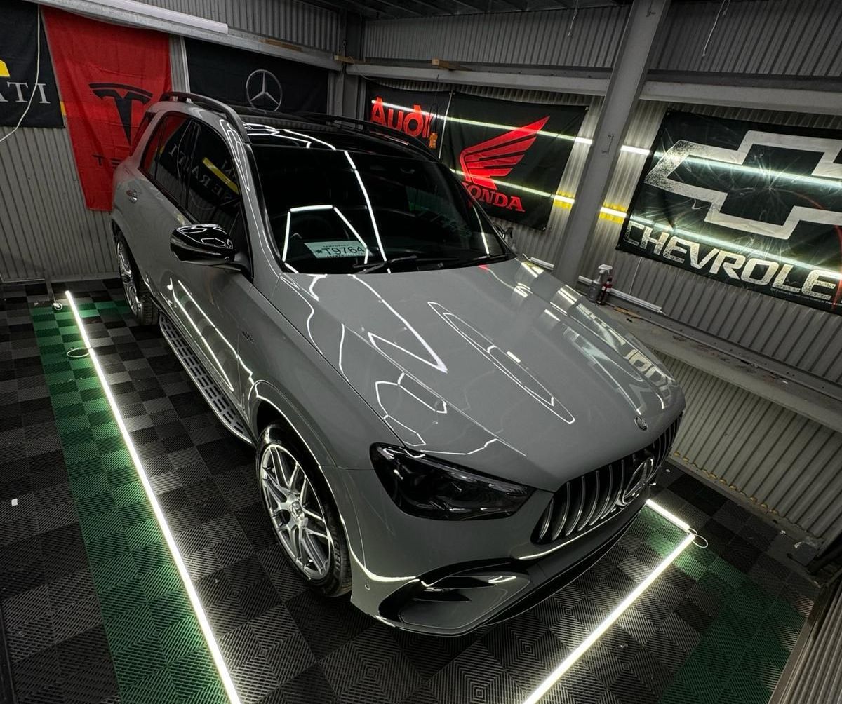 Gray Mercedes SUV parked in a well-lit garage, various car brand flags in the background.