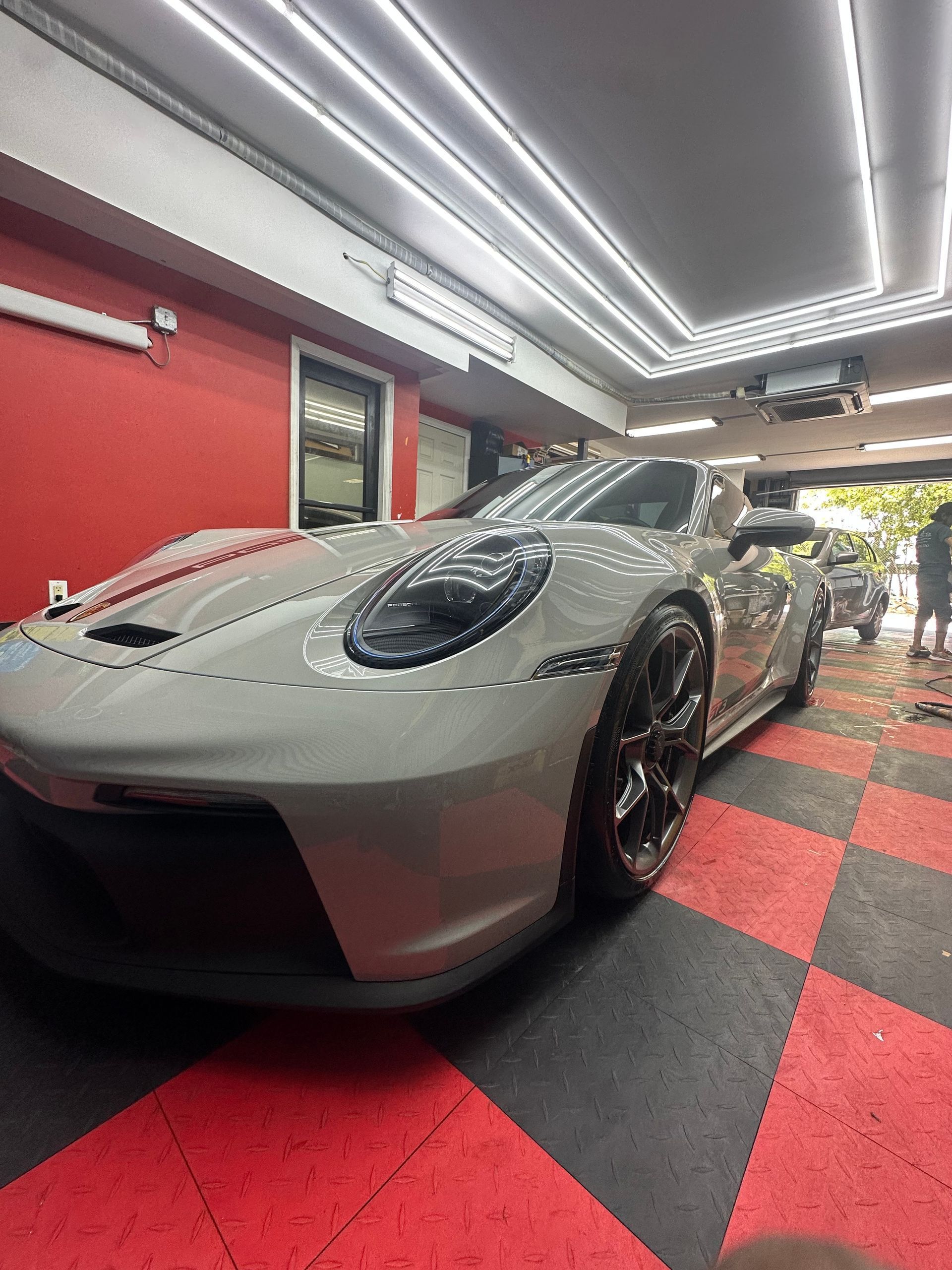 A light gray Porsche sports car inside a garage with red and black checkered flooring and a red wall.