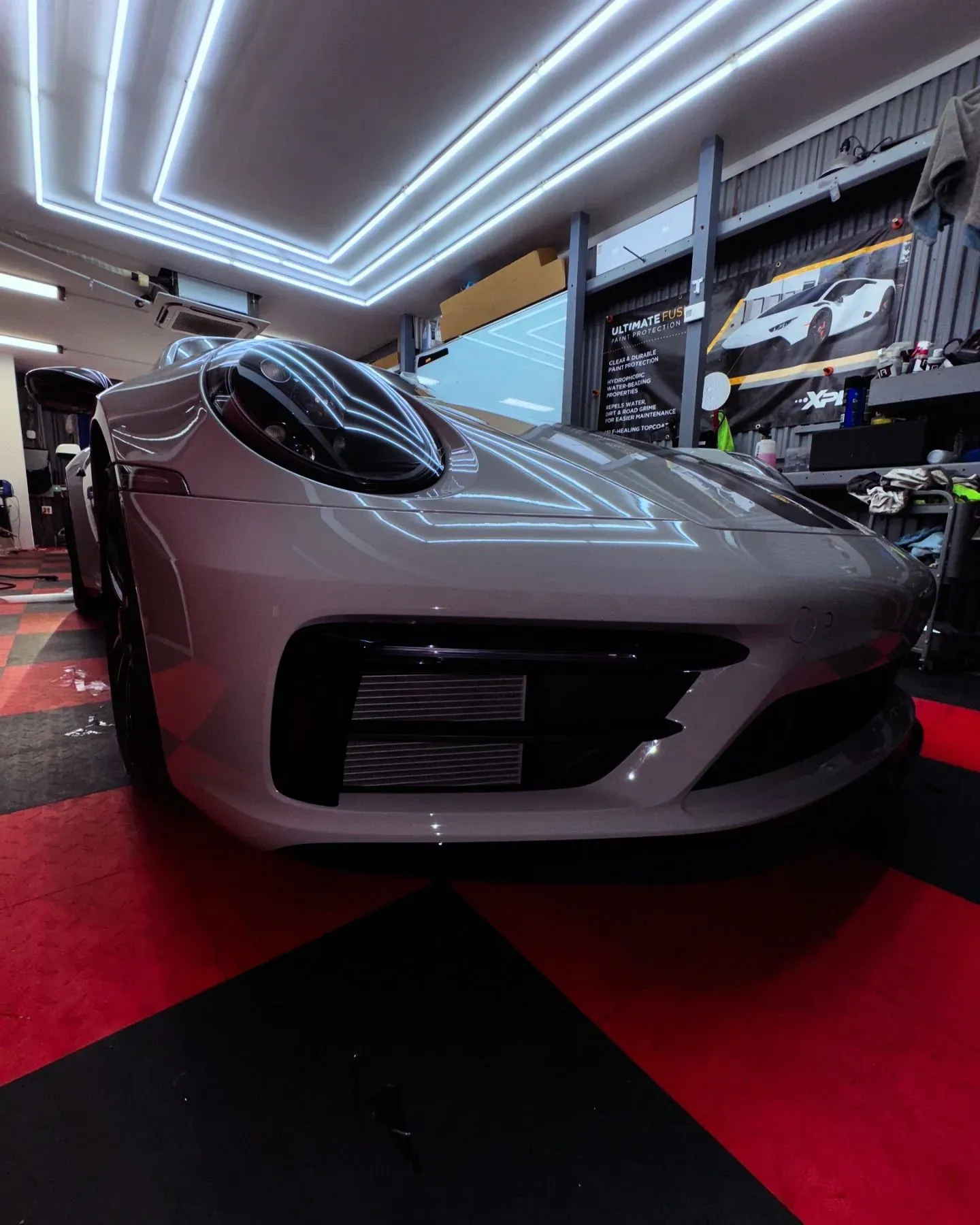 White Porsche 911 parked in a garage with black and red checkered flooring, under bright white lights.