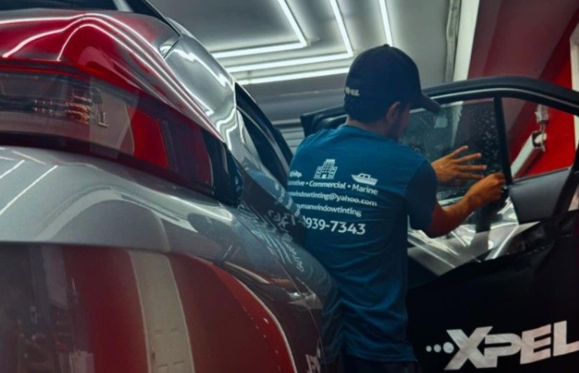 A person in blue shirt installs tinted film on a car window inside a workshop.