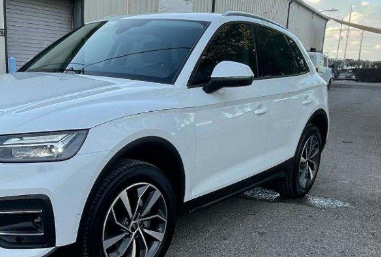 White Audi SUV parked on an asphalt surface. The vehicle is near a building with other cars visible in the background.