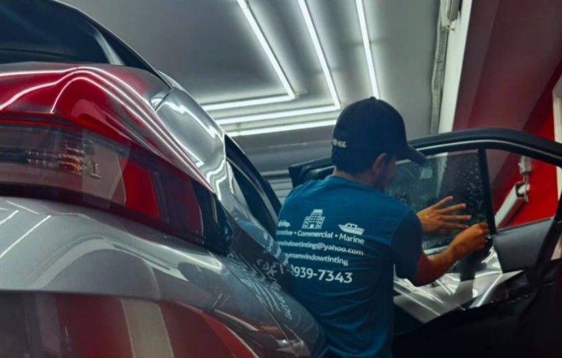 Person installing window tint on a car. The man is wearing a hat and blue shirt.
