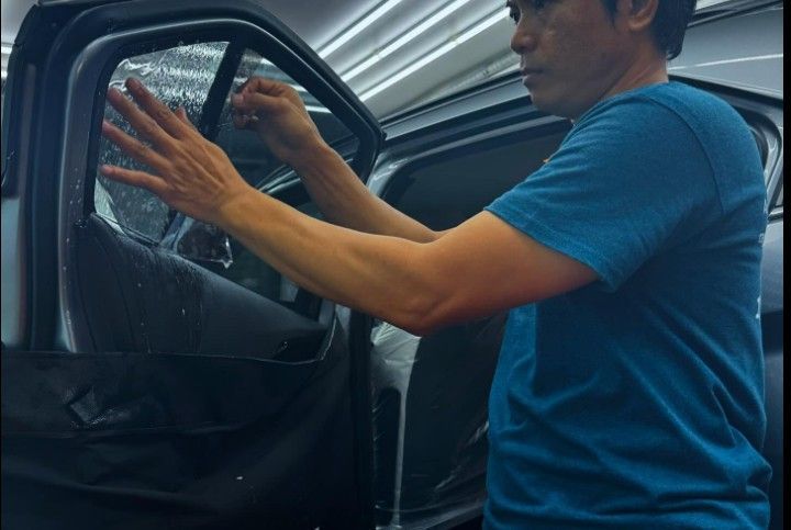 Man applying tint to a car window. He wears a blue shirt and stands indoors near a vehicle.