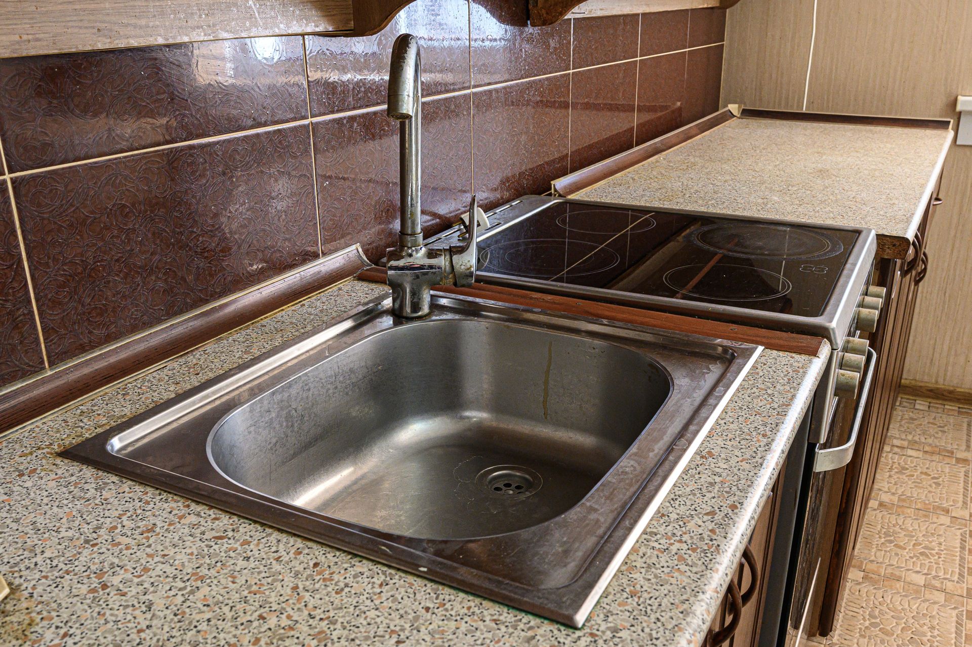 Kitchen sink with chrome faucet, brown tile backsplash, and countertop stove beside it