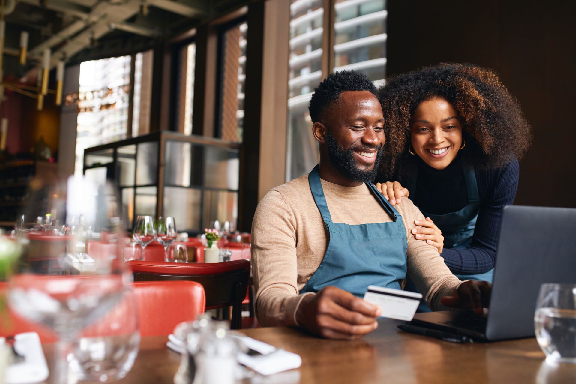 Um homem e uma mulher estão sentados à mesa de um restaurante usando um laptop e um cartão de crédito.