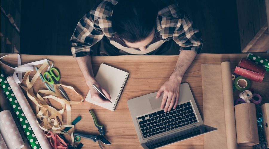 Um homem está sentado em uma mesa com um laptop e escrevendo em um caderno.