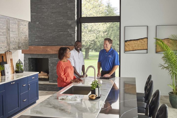 Three people standing in a modern kitchen by a large island and fireplace, talking.
