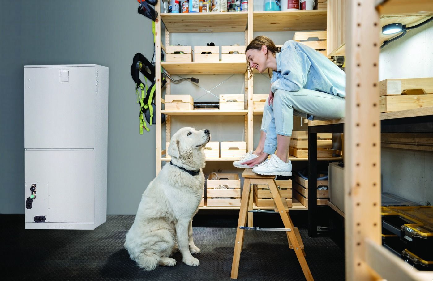 Woman sitting on a stool with a white dog in a home office beside shelves and a refrigerator