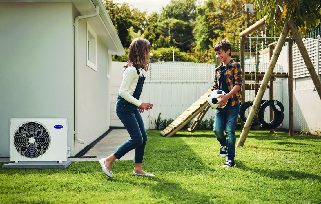 Two people play catch in a sunny backyard beside a house and outdoor AC unit.