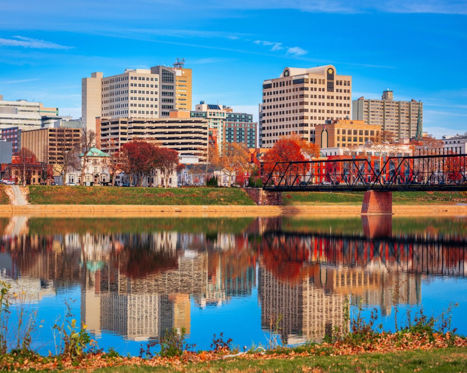 City skyline and red bridge reflected in a calm river under a clear blue sky.