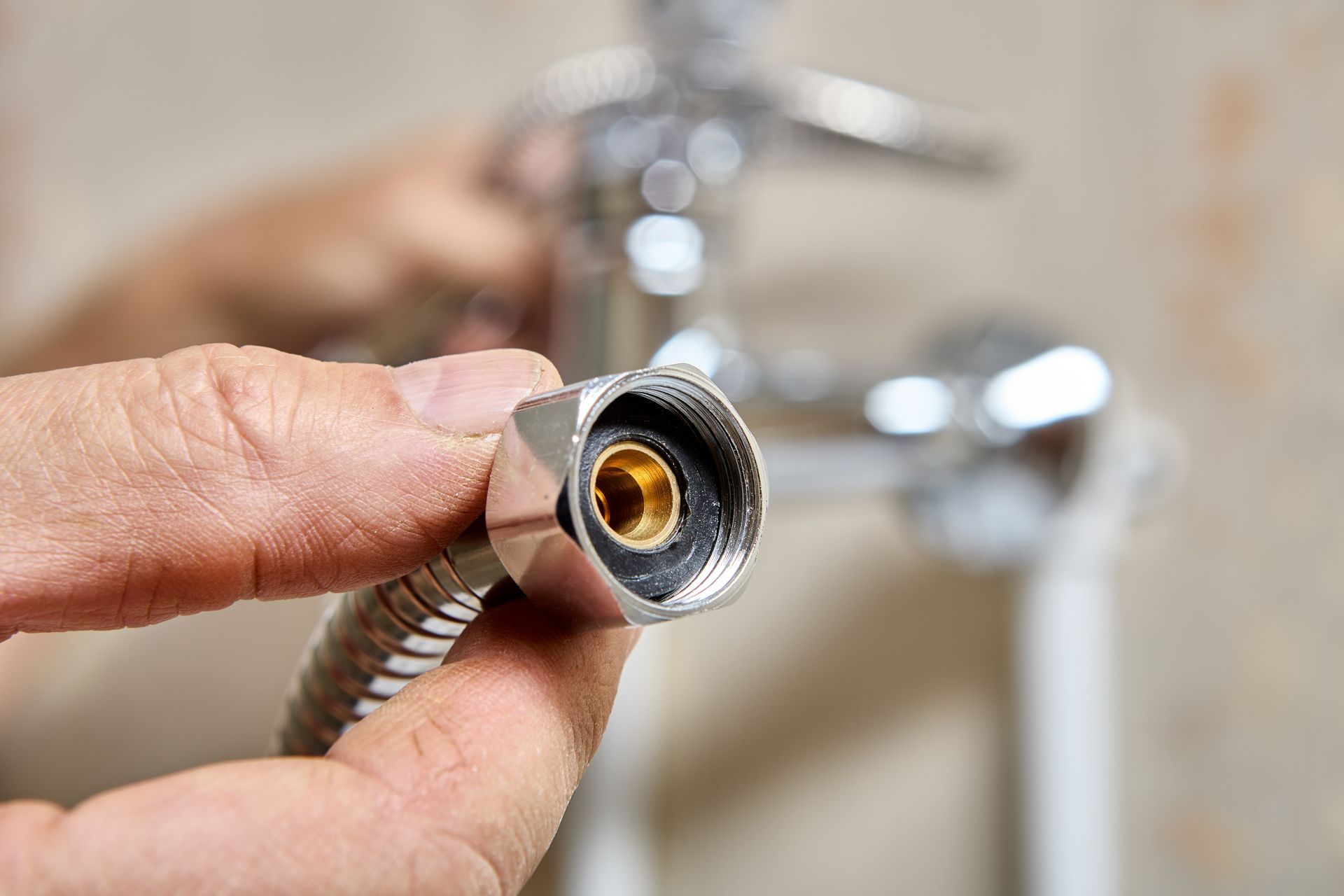 Hand holding a braided faucet hose connector near a blurred sink faucet, showing the threaded metal end.