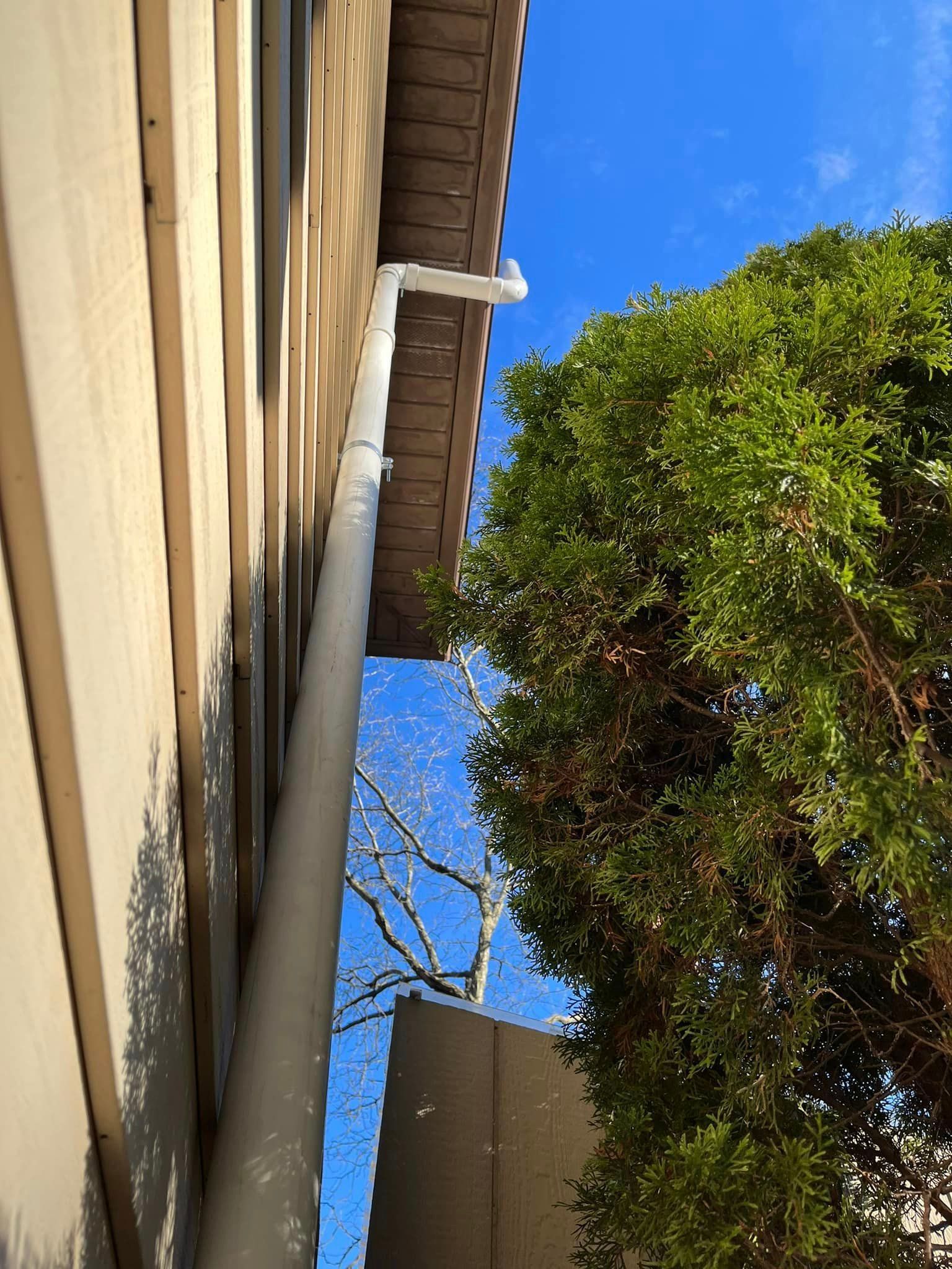 A white pipe is hanging from the side of a building next to a tree.