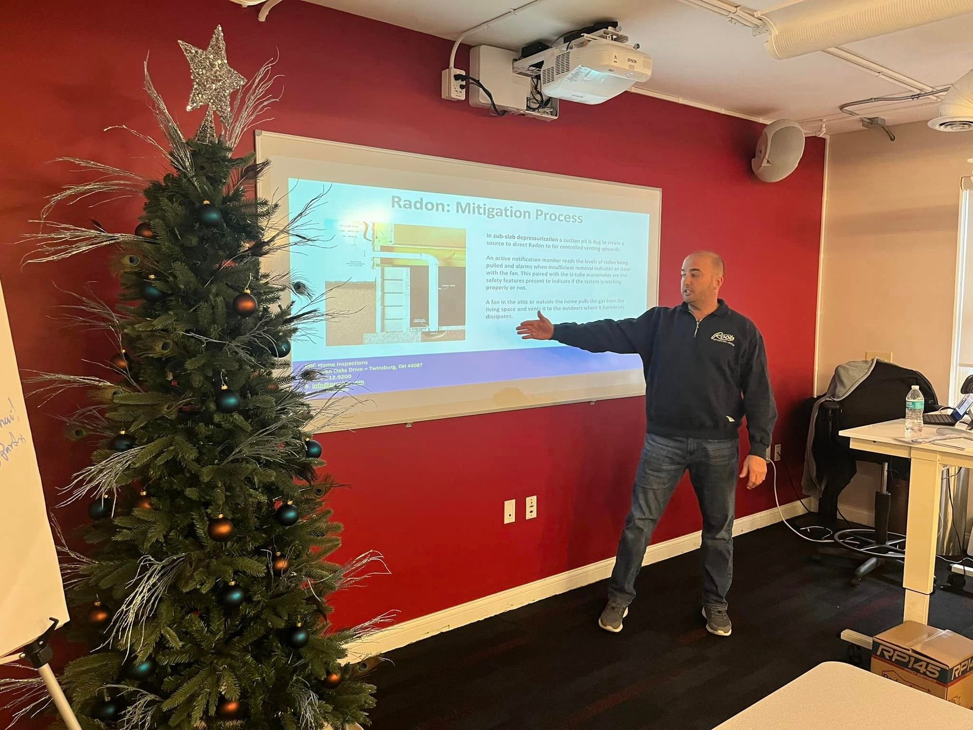 A man is standing in front of a projector screen in a room with a christmas tree.