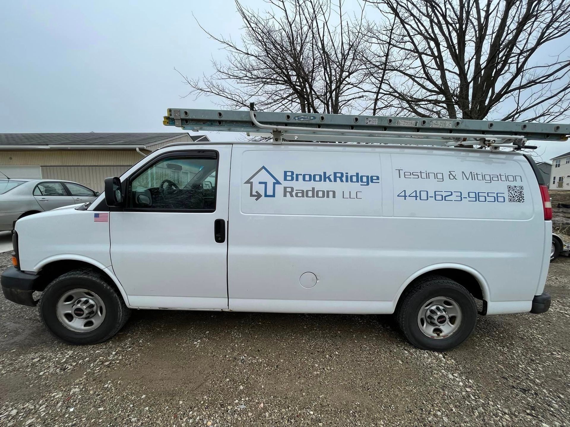 A white van with a ladder on top of it is parked in a gravel lot.