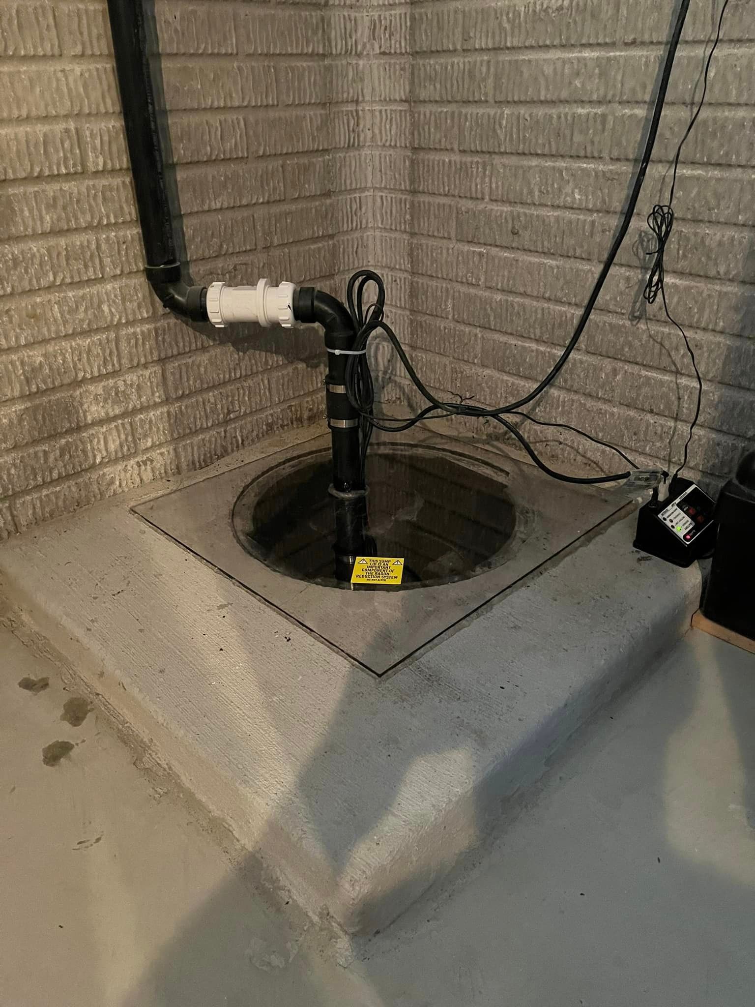 A sump pump is sitting in a corner of a basement next to a brick wall.