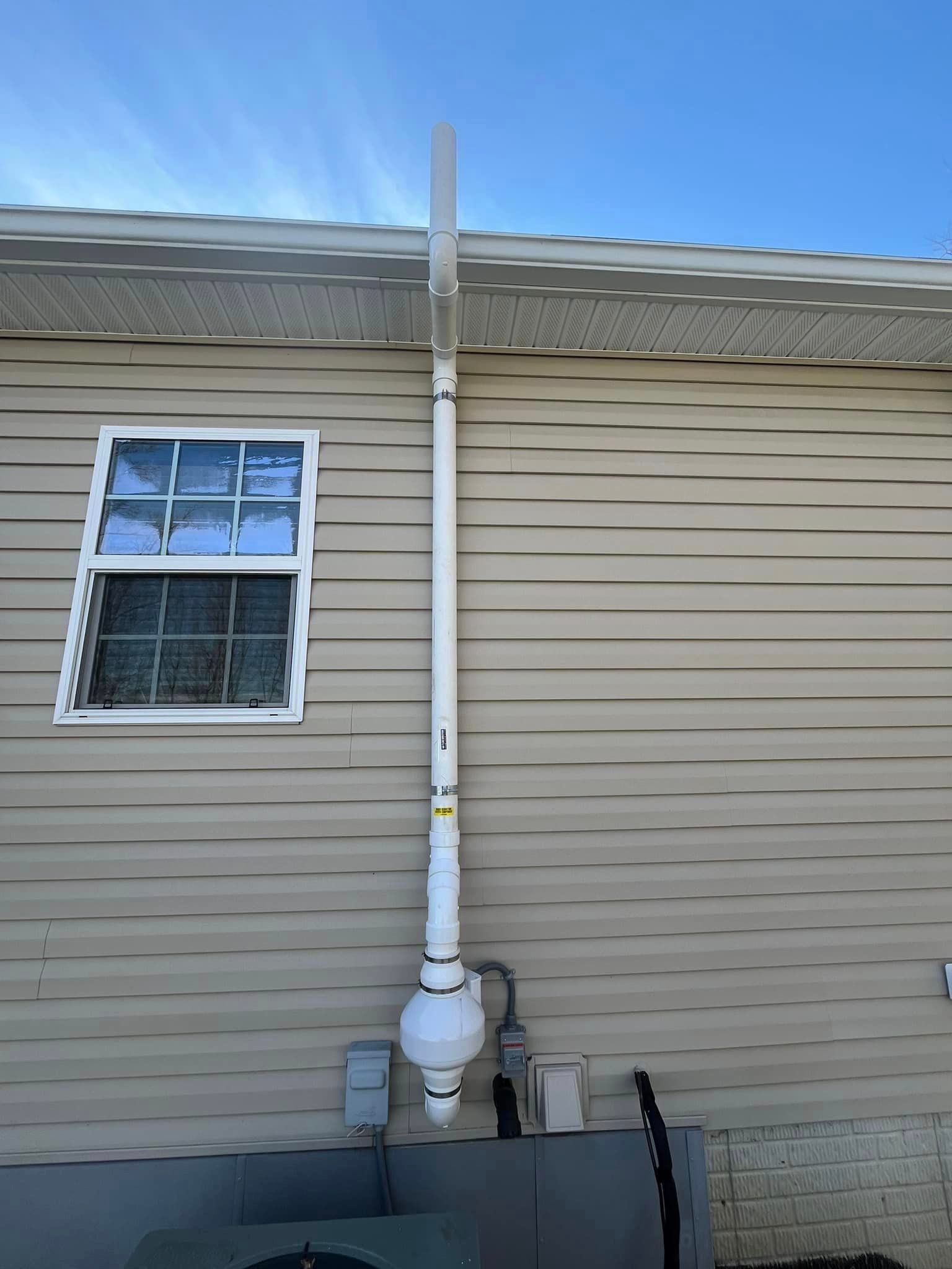 A white pipe is attached to the side of a house next to a window.