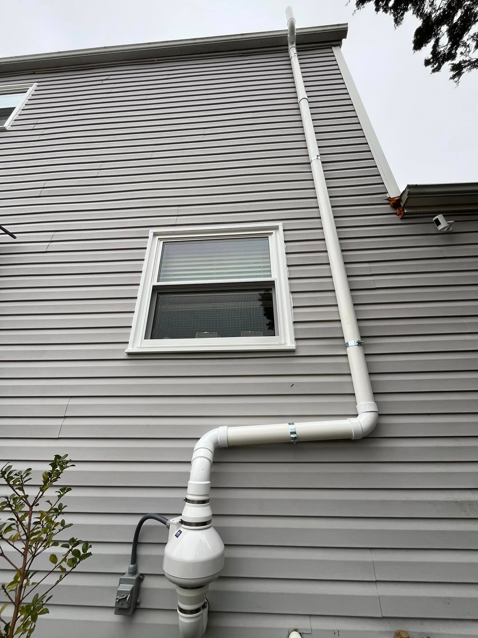 A white pipe is attached to the side of a house next to a window.