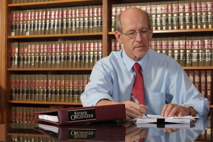 A man in a blue shirt and red tie is sitting at a desk in front of a bookshelf.