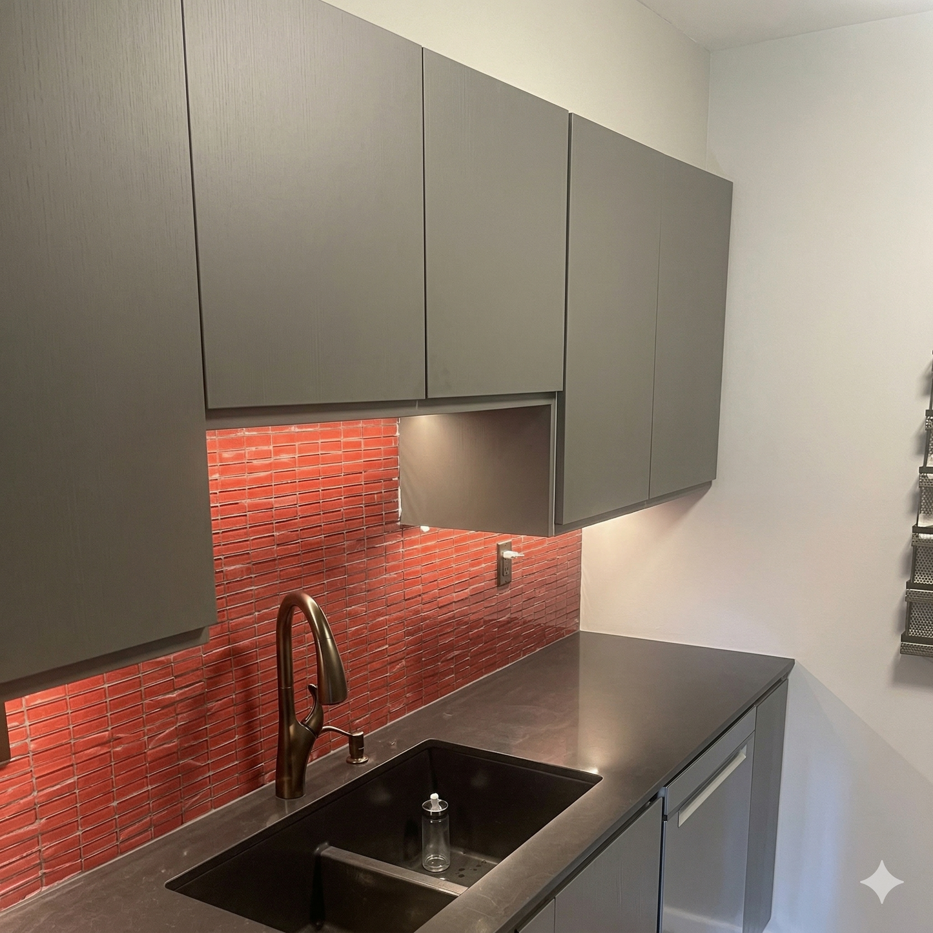 A kitchen area with grey flat-panel cabinets, a red glass tile backsplash, a dark sink, and a modern gold faucet.