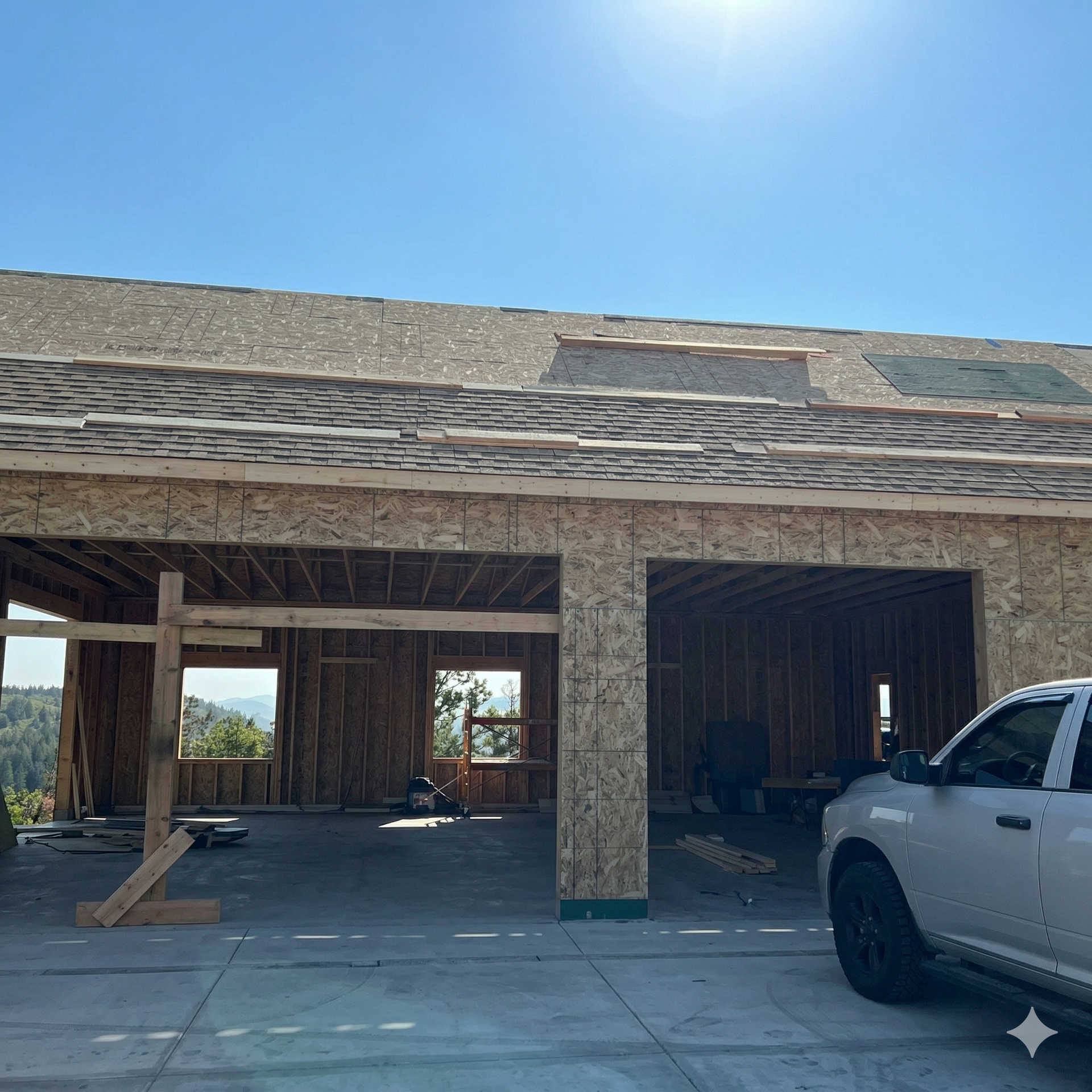 A partially framed, open-concept garage structure with a shingled roof under a bright blue sky, with a truck parked nearby.
