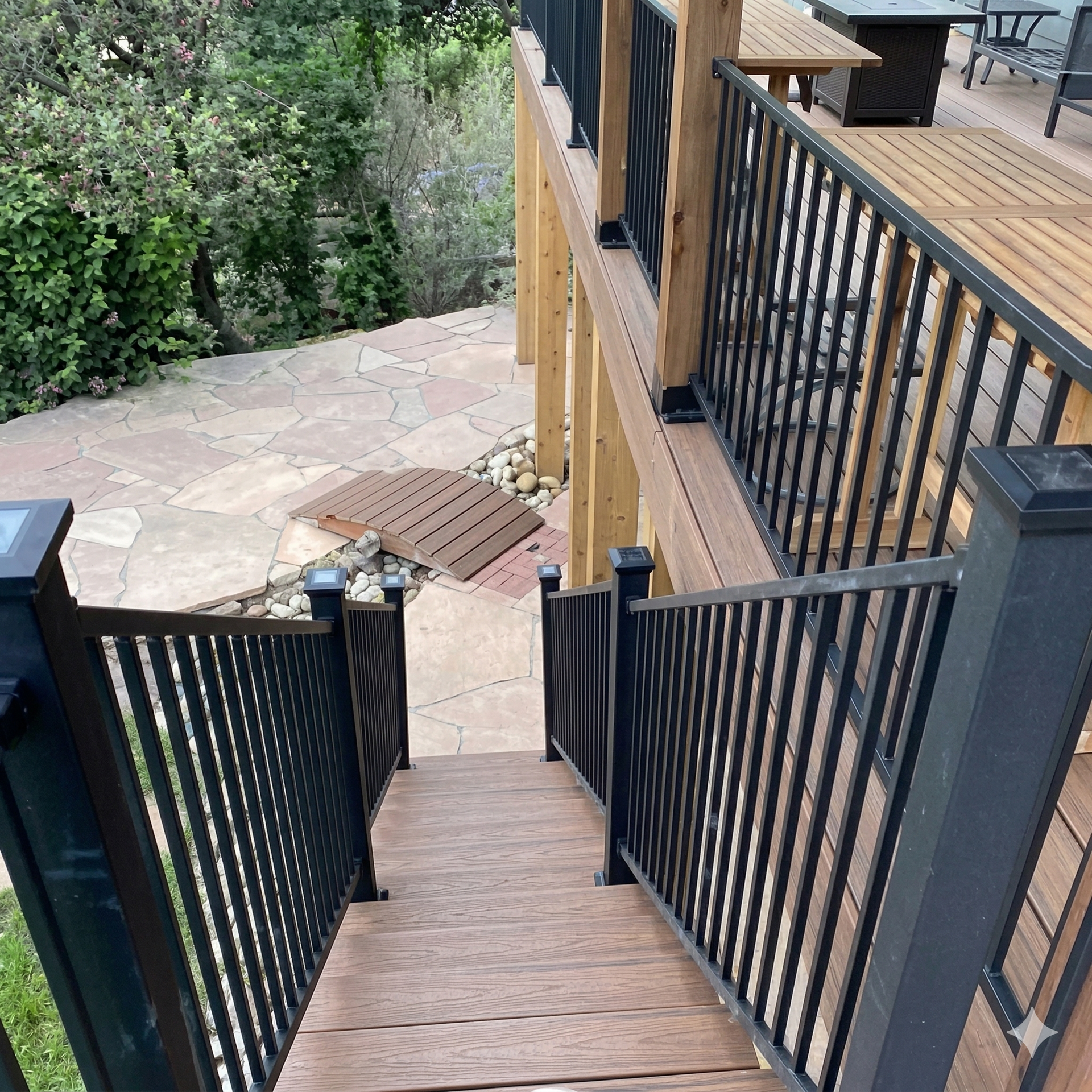 A view from the top of wooden stairs leading down to a stone patio, framed by dark metal railings on both sides.