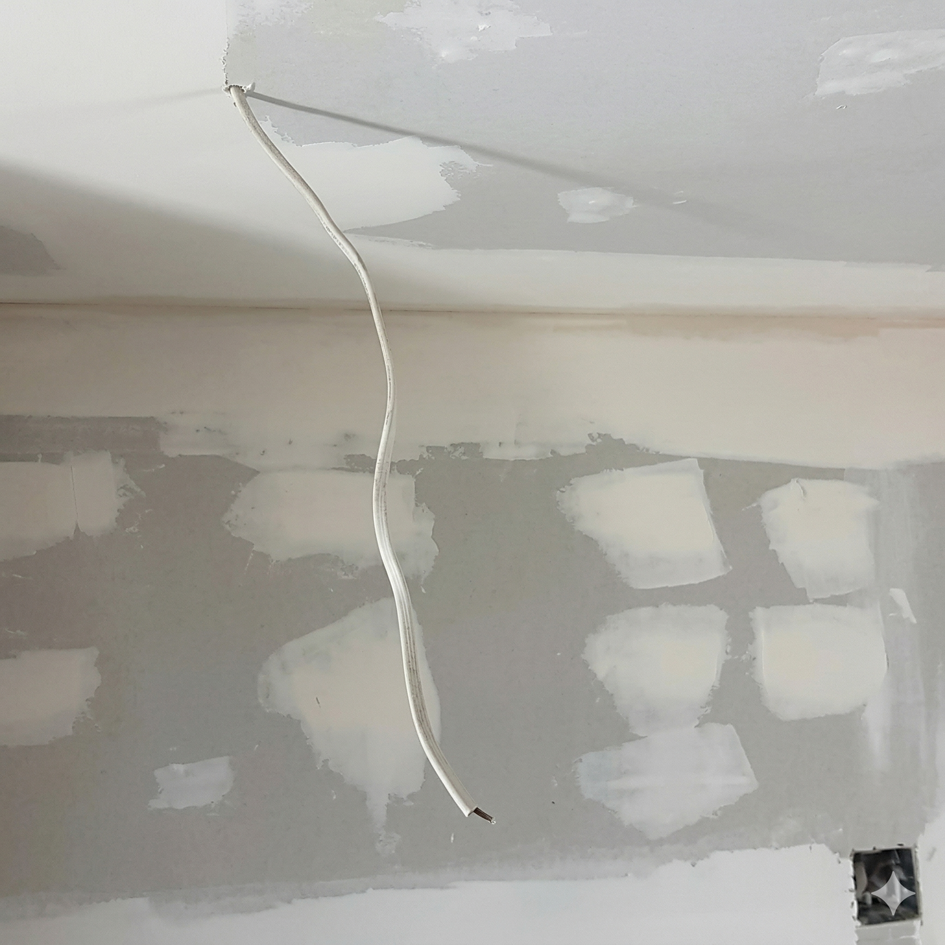 A white electrical wire hangs from an unfinished drywall ceiling above a wall with plastered joints and nail heads.