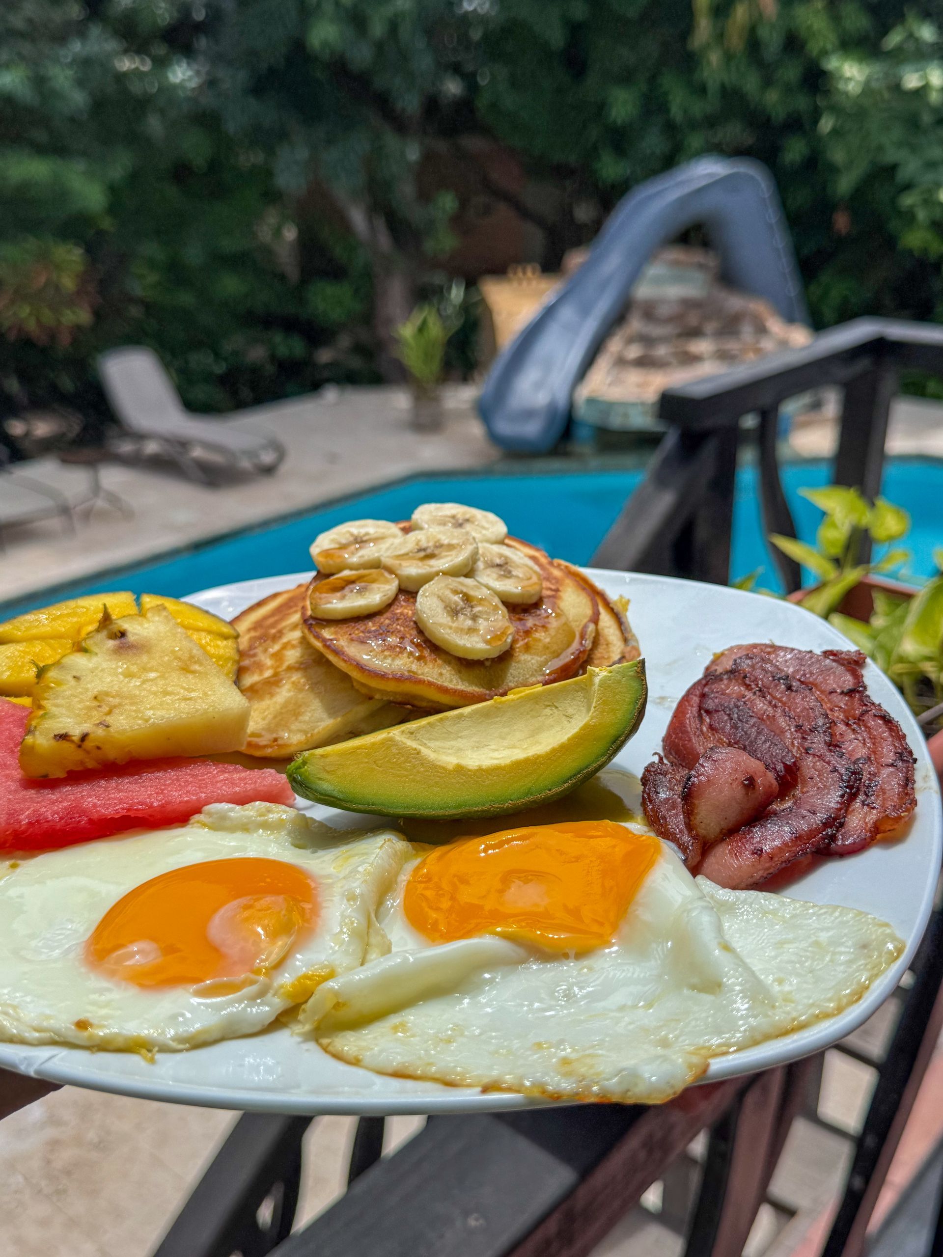 Breakfast plate with eggs, avocado, pastries, and bacon by a poolside table at Caribbean Shores Bed and Breakfast Belize