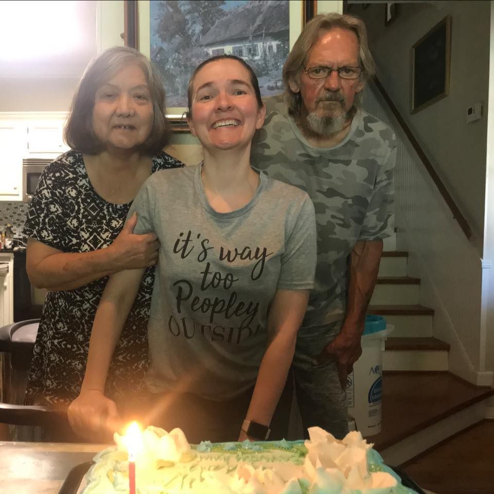 Three people smiling behind a birthday cake with lit candles in a home staircase setting