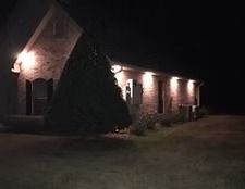 Brick house at night with lit exterior lamps and a large evergreen tree in front