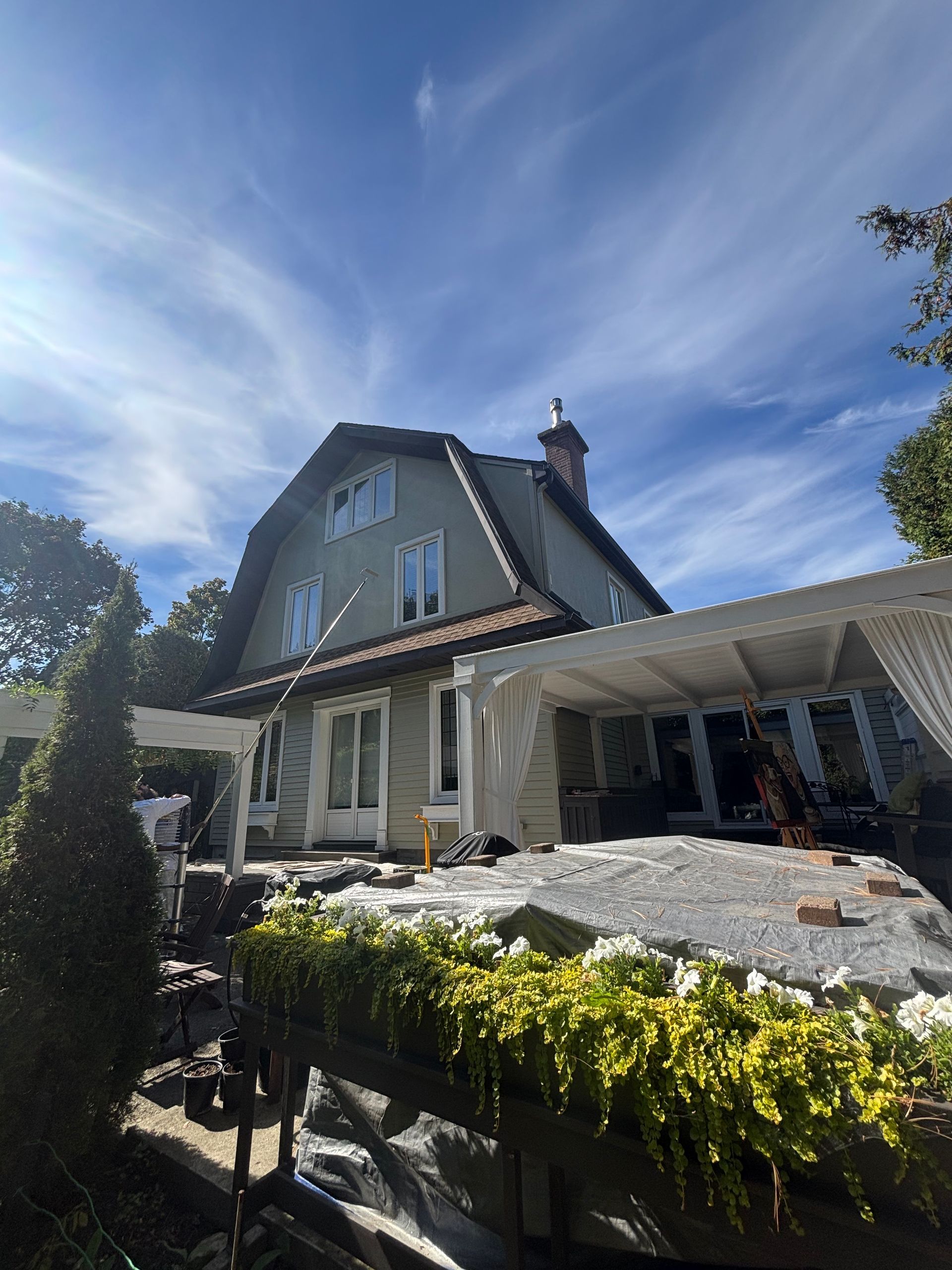 Maison ensoleillée au toit pentu et patio couvert, entourée de verdure sous un ciel bleu.