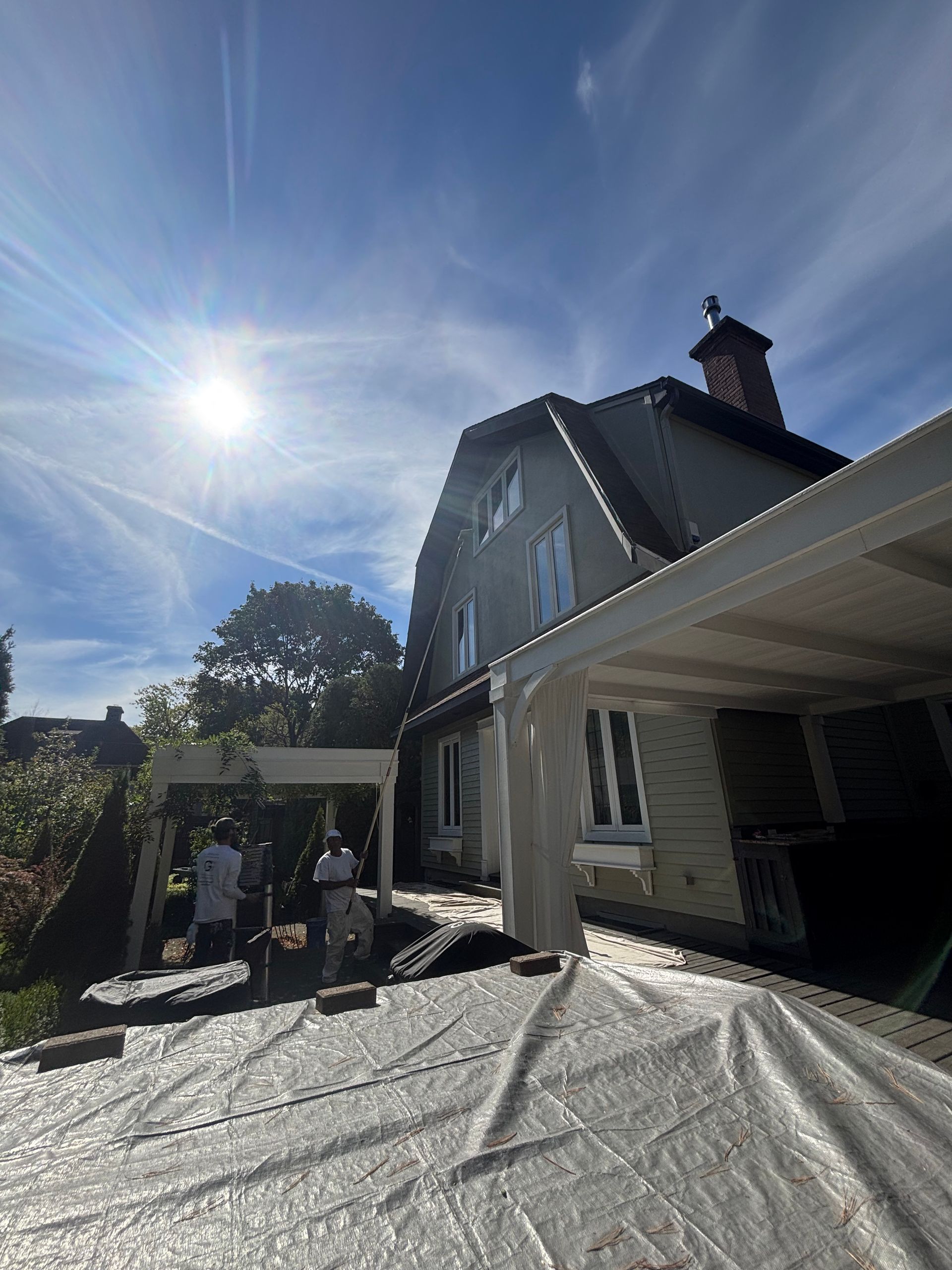 Extérieur d'une maison baigné de soleil, avec des personnes sur une terrasse recouverte d'une bâche sous un auvent blanc.