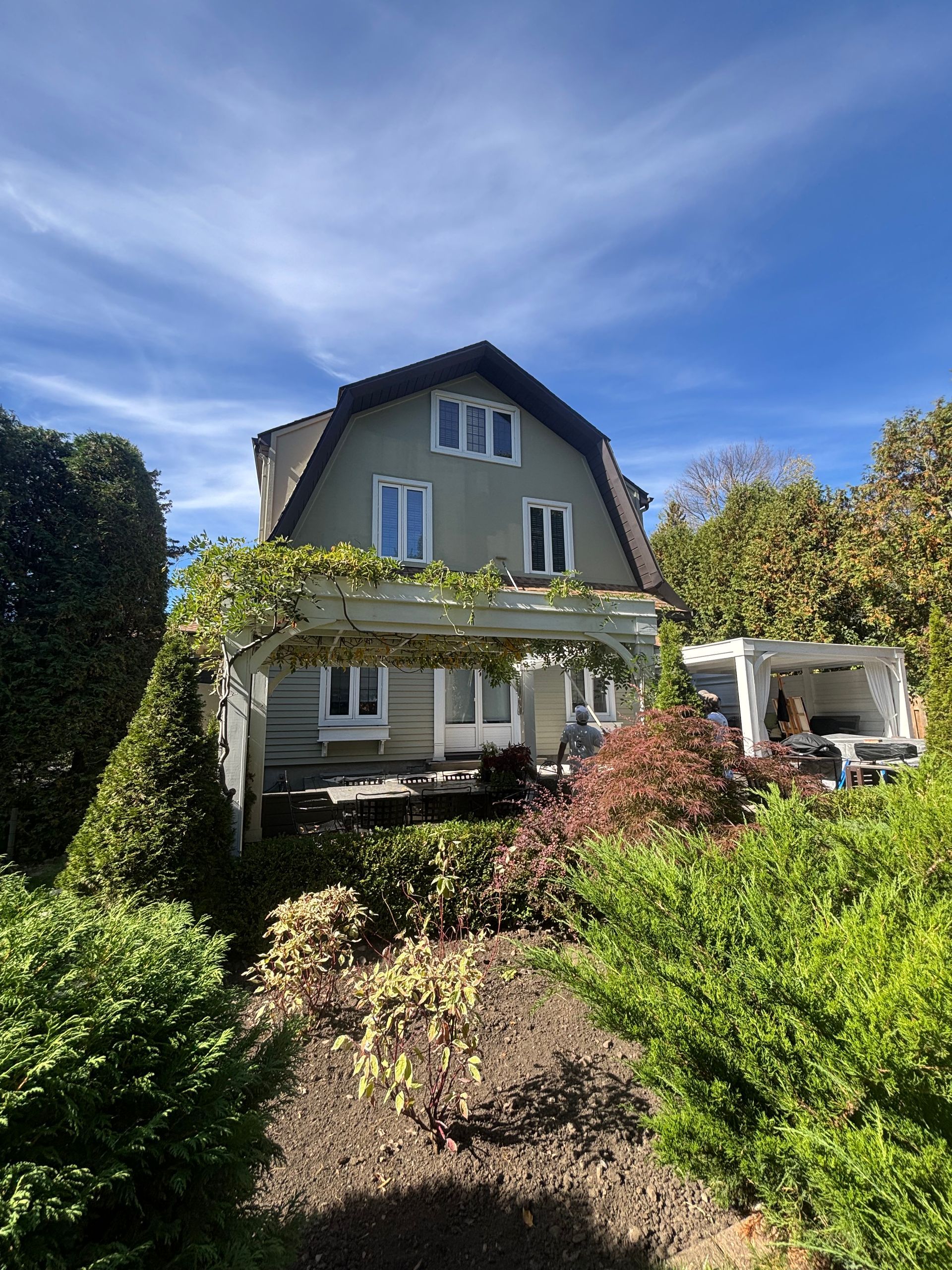 Maison blanche avec porche recouvert de lierre, entourée d'un jardin luxuriant sous un ciel d'un bleu éclatant.