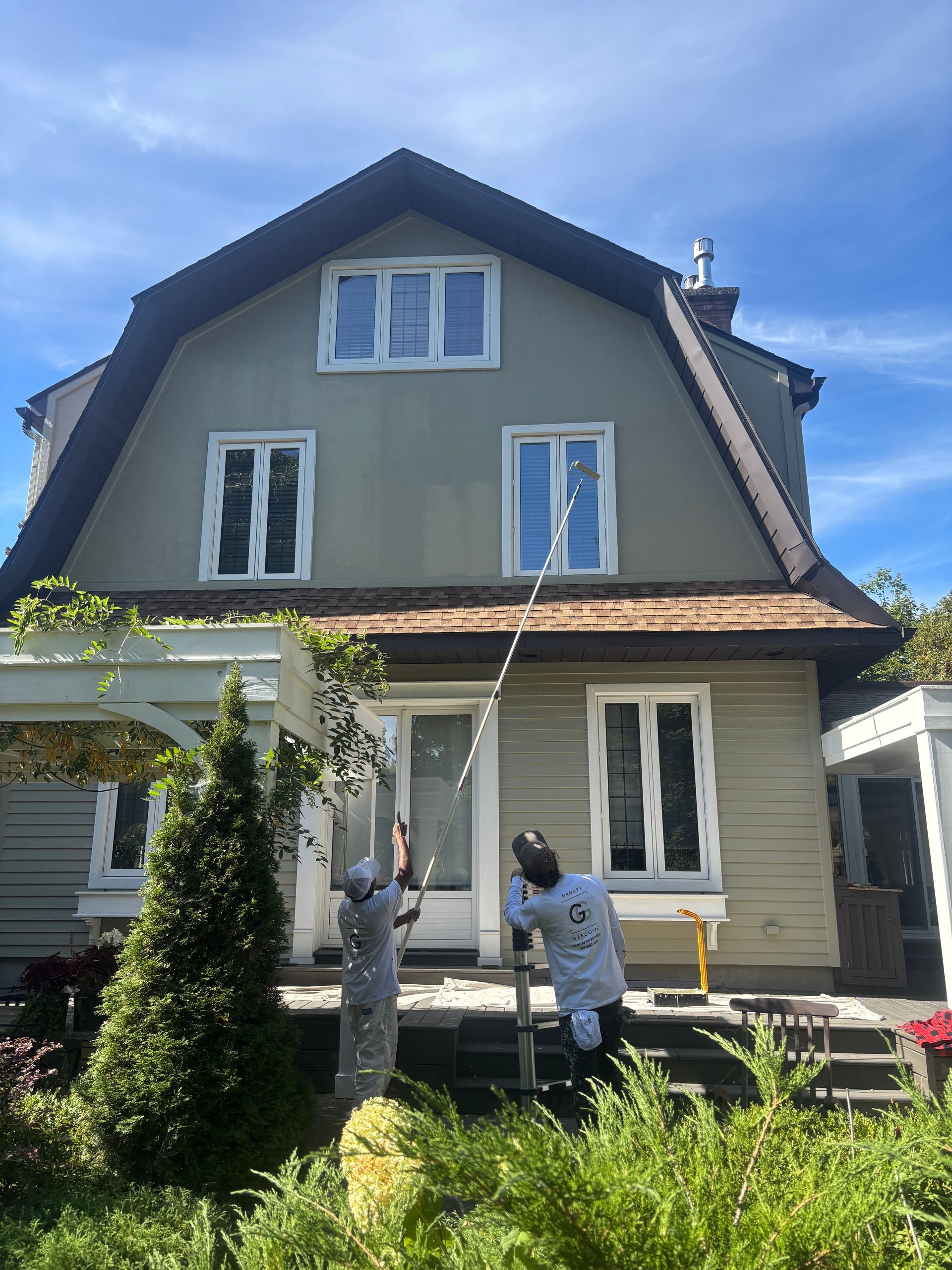 Deux personnes peignent le porche d'une maison grise au toit foncé et aux boiseries blanches.