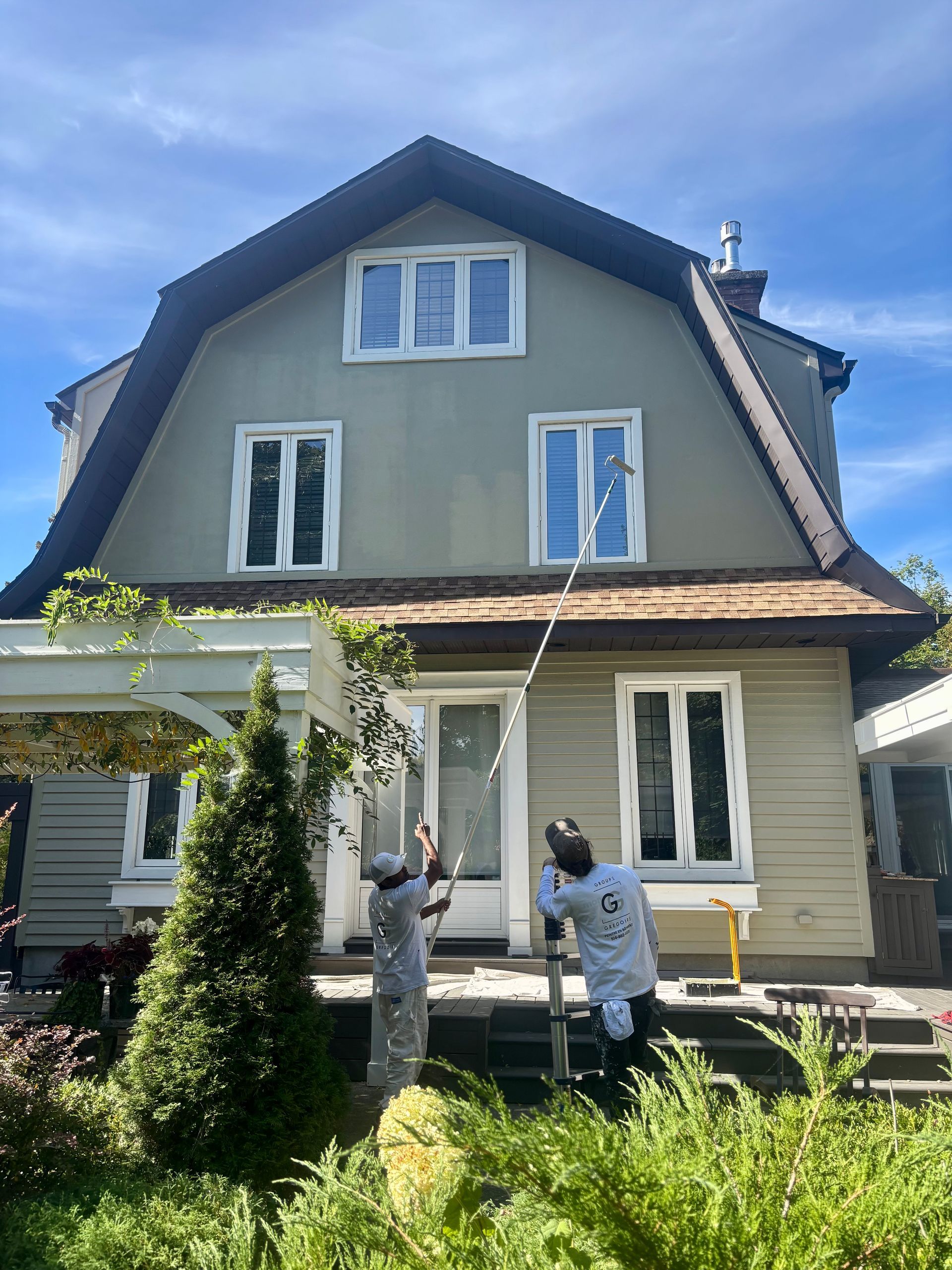 Deux personnes peignent la façade supérieure d'une maison sous un ciel d'un bleu éclatant.