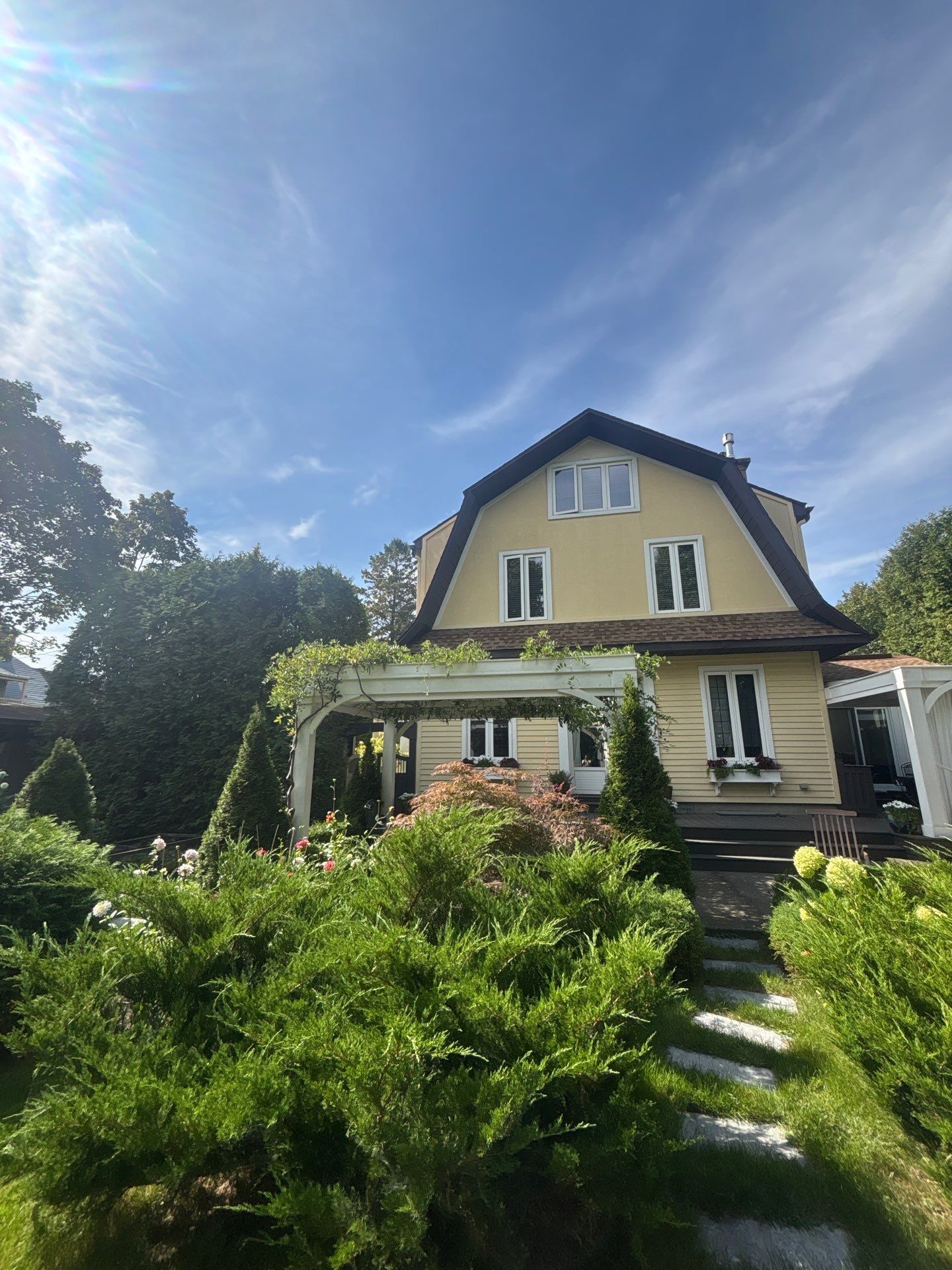 Maison jaune aux boiseries blanches et à la véranda, entourée d'une végétation luxuriante sous un ciel bleu.