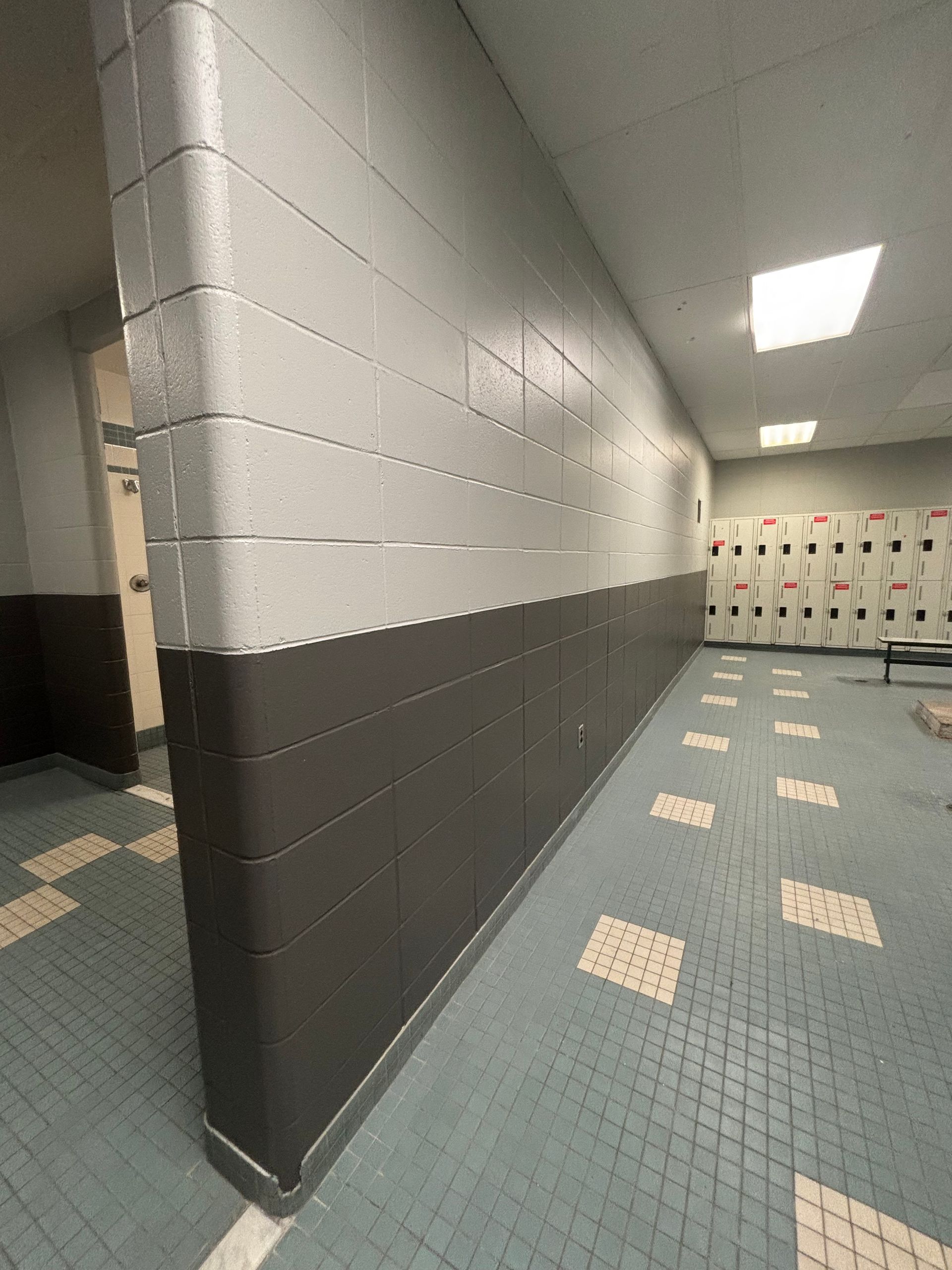 Couloir d'école aux murs en parpaings gris et blancs et à la moquette bleue à motifs, menant à des casiers ou des armoires.