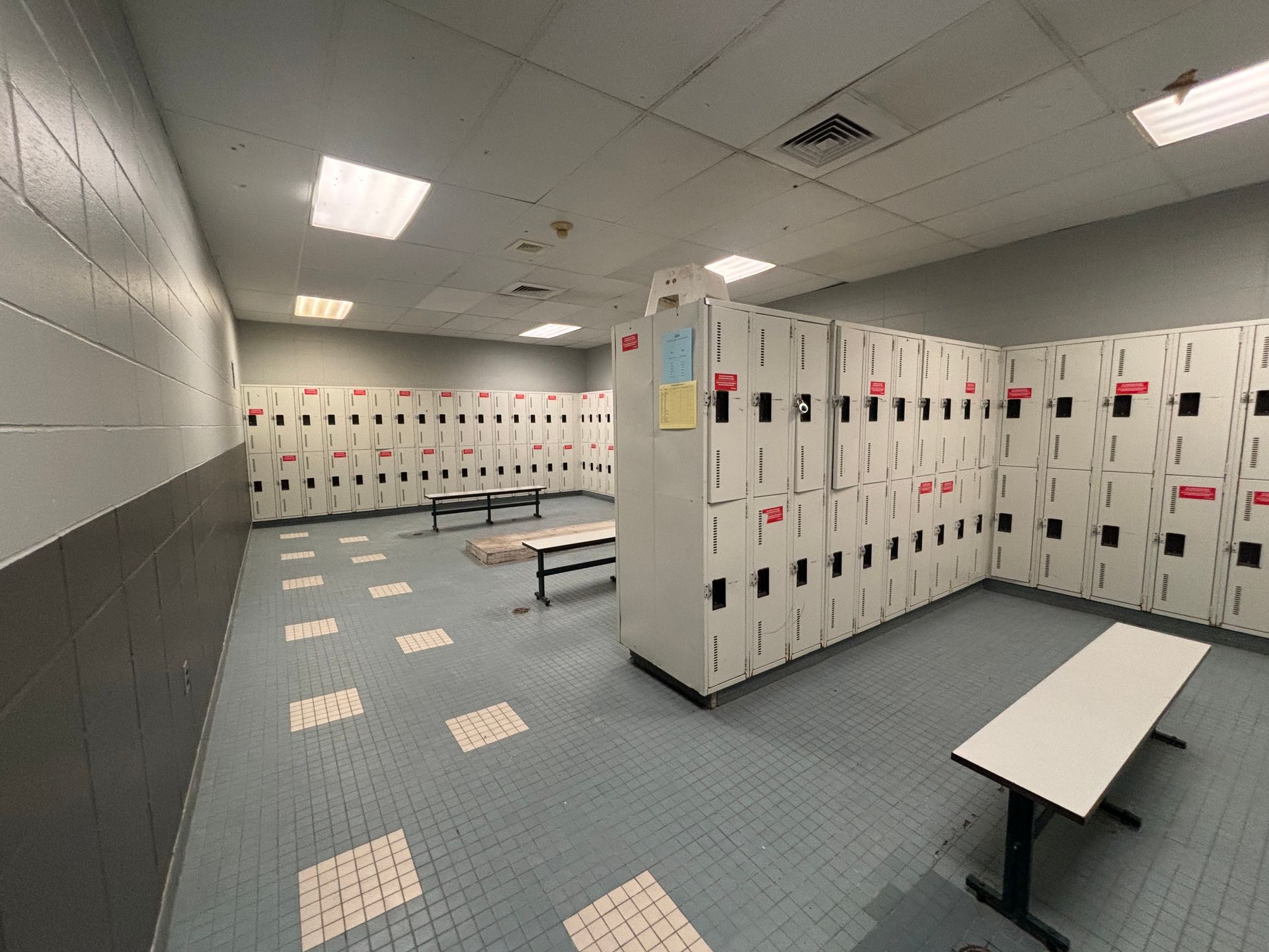 Vestiaire d'école vide avec casiers blancs, sol carrelé bleu et bancs sous des néons