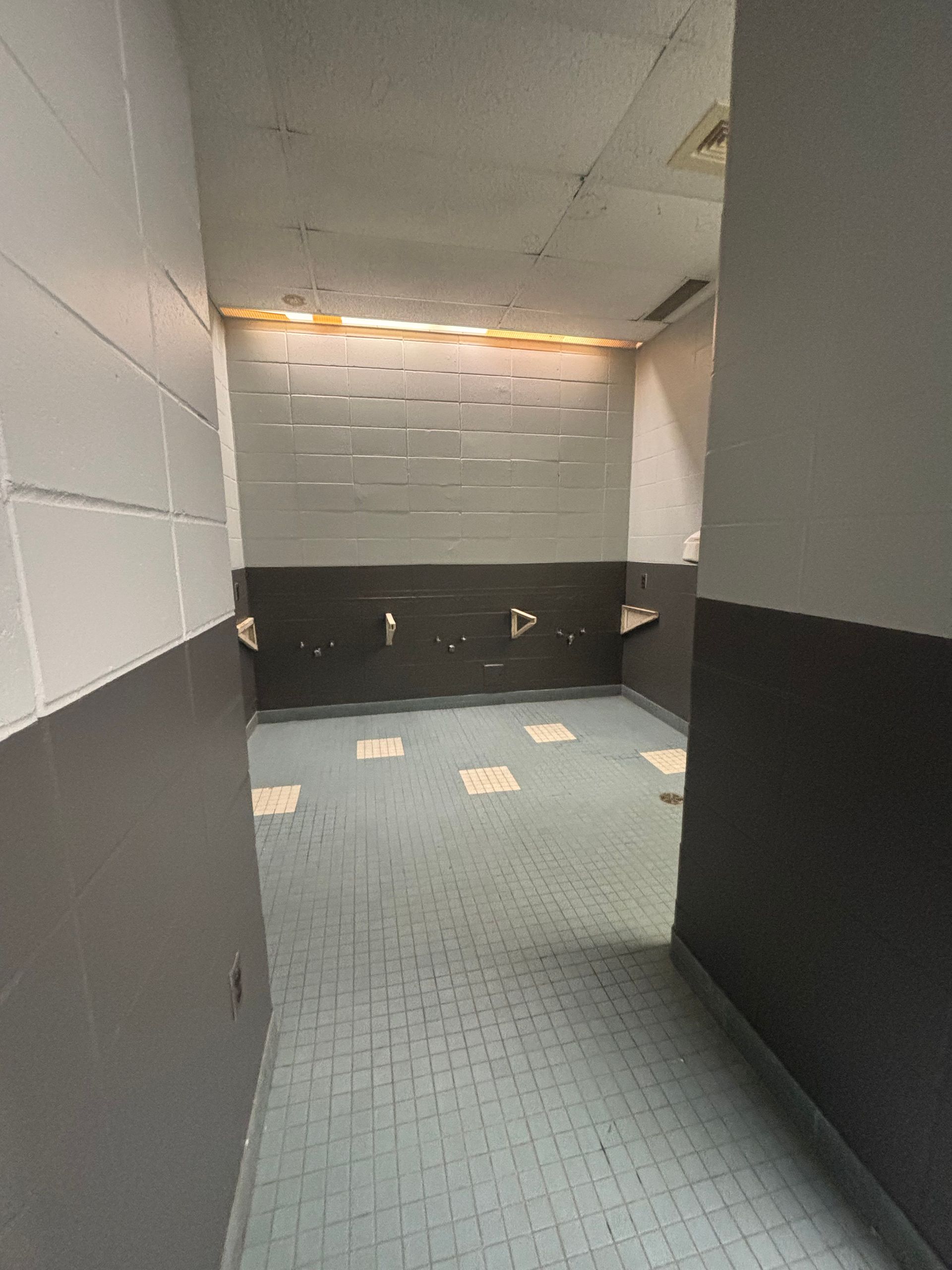 Toilettes vides avec murs carrelés gris et blancs, sol gris et une rangée de lavabos sous une lumière chaude.