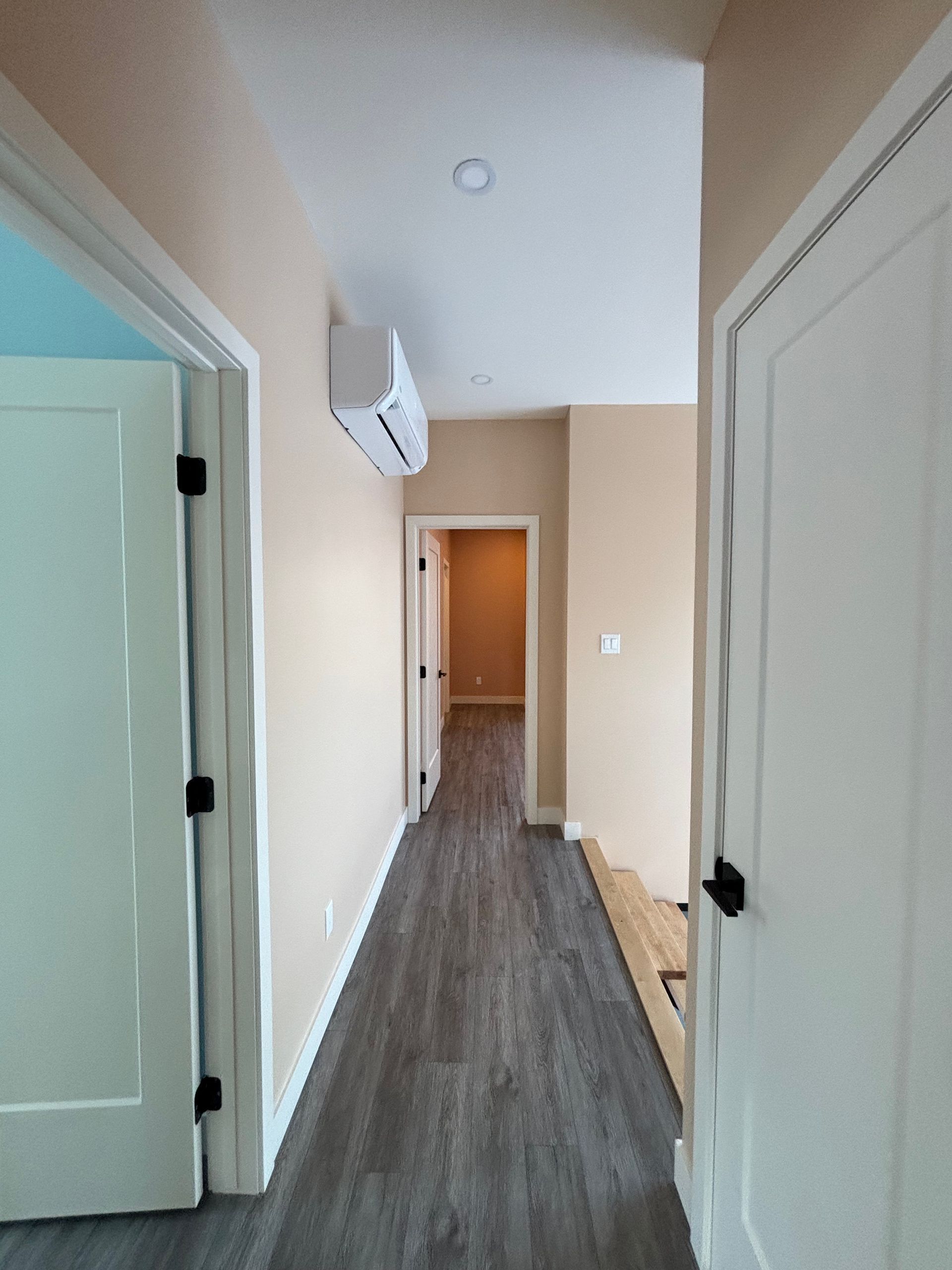 Couloir étroit avec portes blanches, murs beiges et parquet gris menant à une pièce éclairée.
