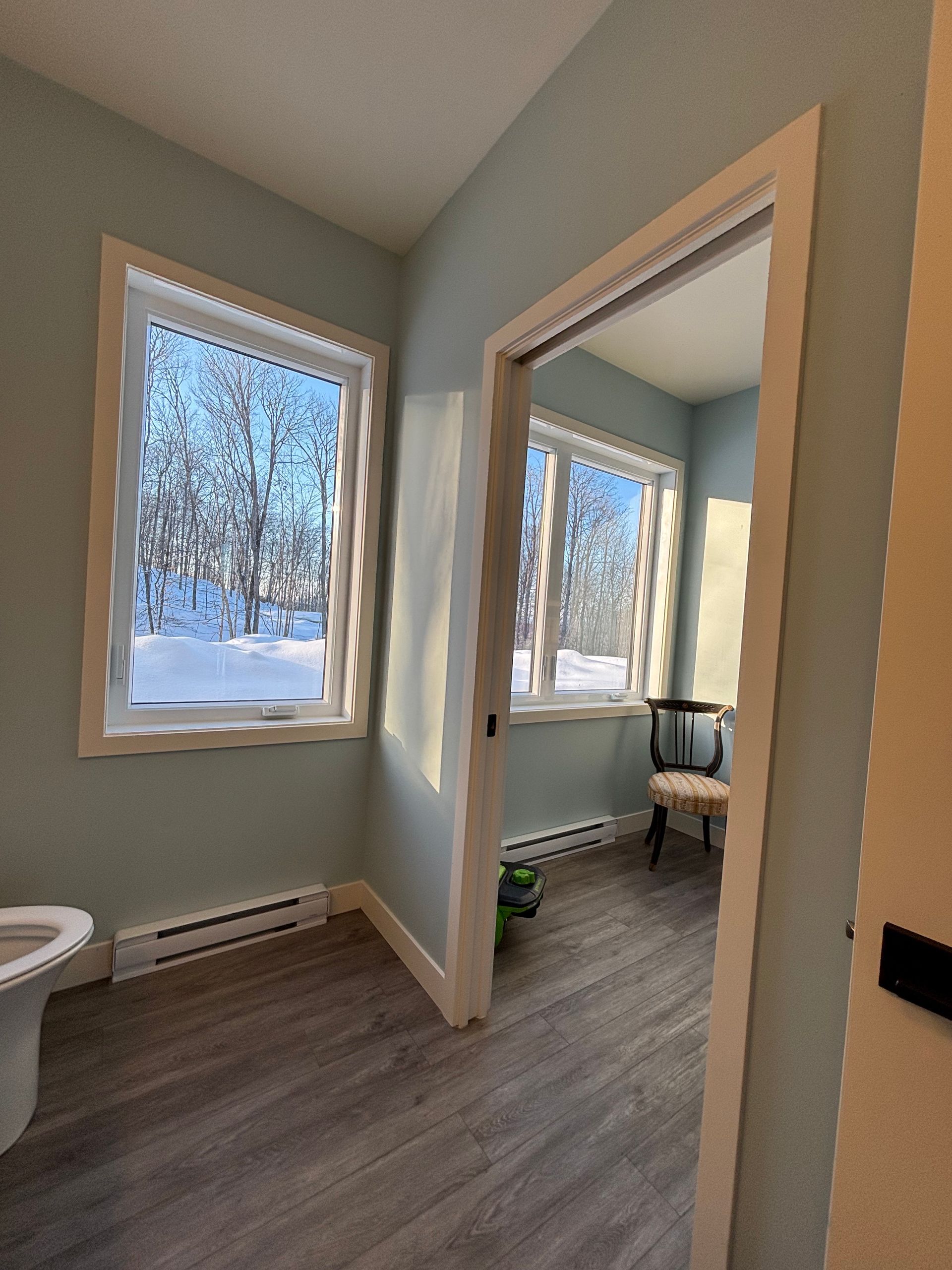 Petite salle de bains avec toilettes, deux fenêtres et vue sur une chambre donnant sur un paysage enneigé.