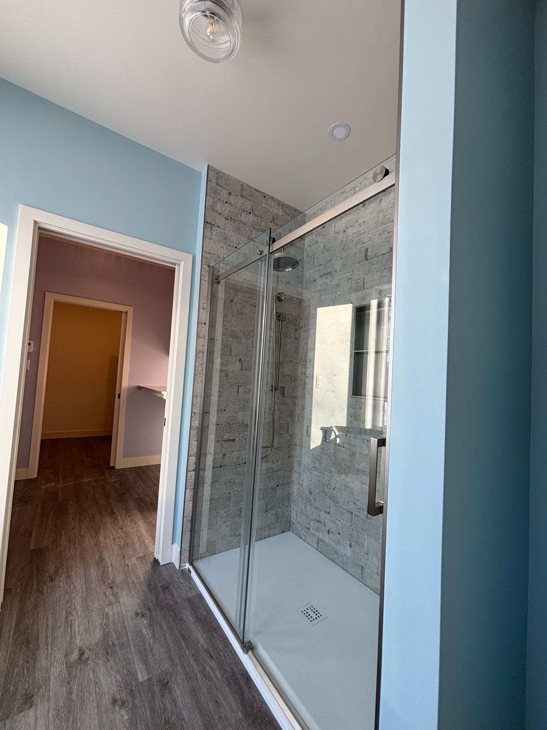 Salle de bains moderne avec douche en verre, carrelage gris et murs bleus donnant sur une porte ouverte