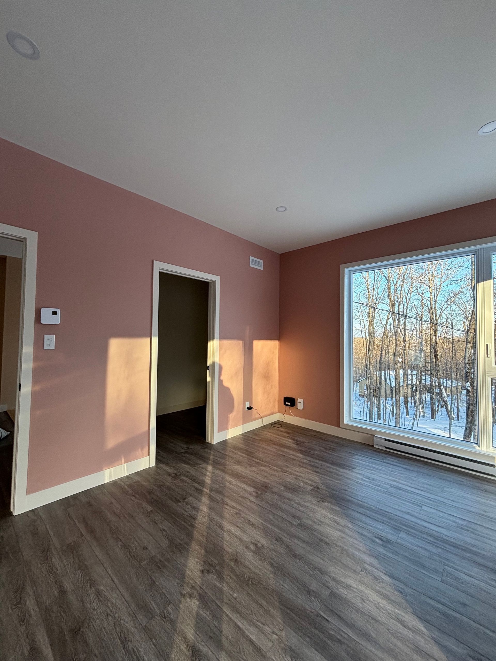 Pièce vide aux murs roses, au parquet foncé et dotée d'une grande fenêtre laissant entrer la lumière du soleil.