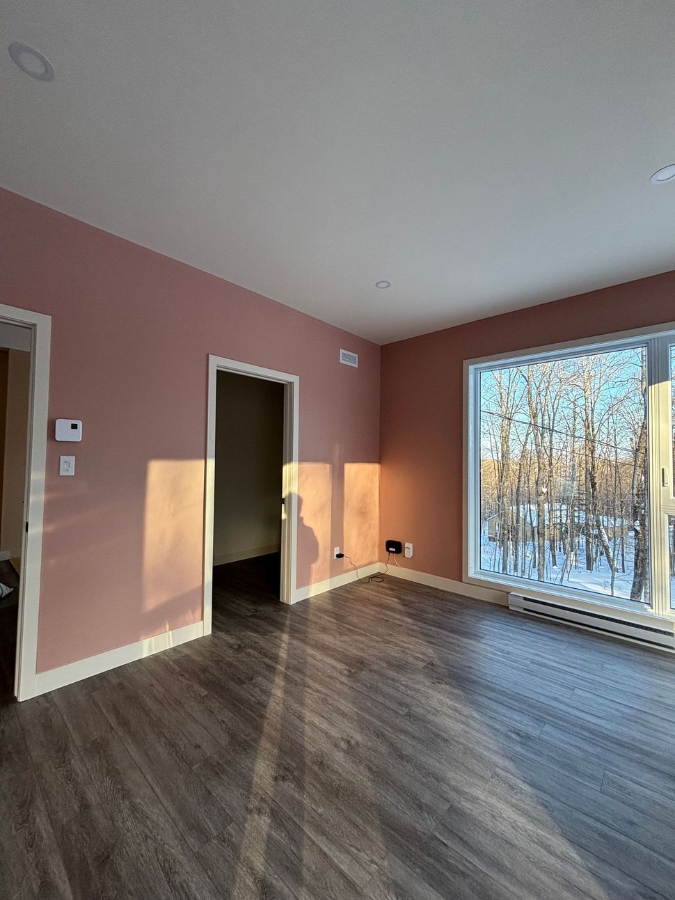 Pièce rose vide avec parquet foncé et grande fenêtre laissant entrer la lumière du soleil