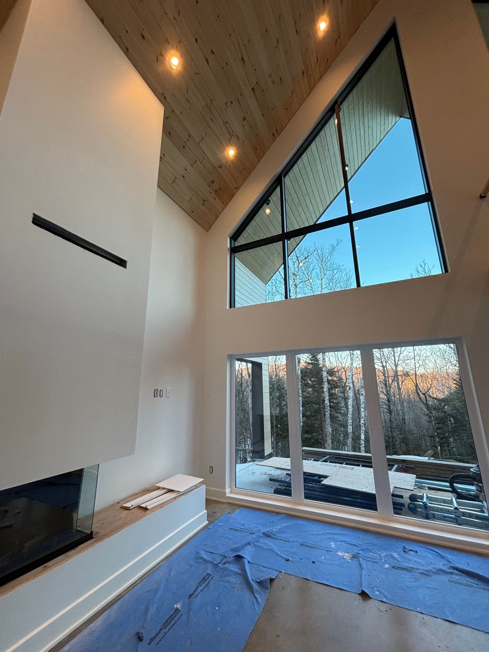 Salon moderne avec plafond voûté en bois, grande fenêtre triangulaire et canapé recouvert de tissu bleu face à une cheminée.