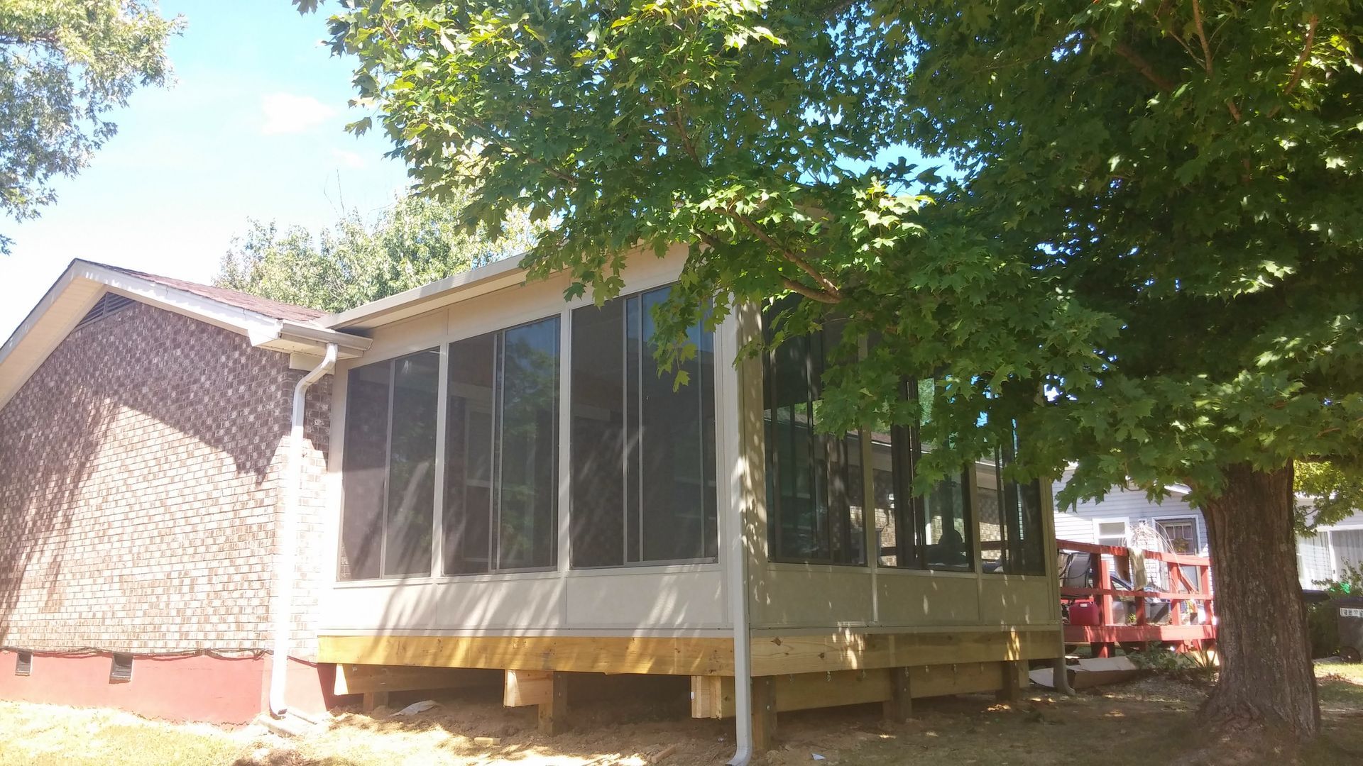 A sunroom with large windows and a wooden base built onto the side of a brick house, partially shaded by a large tree.