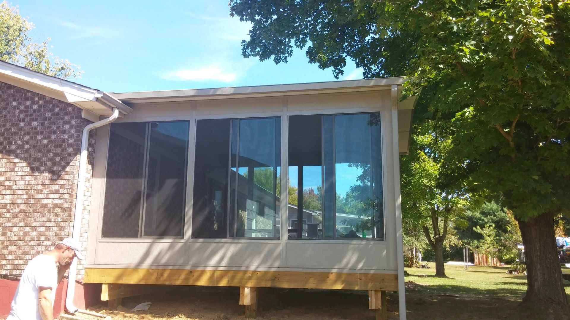 A person standing outside a newly constructed sunroom addition with glass panels and a wooden frame.