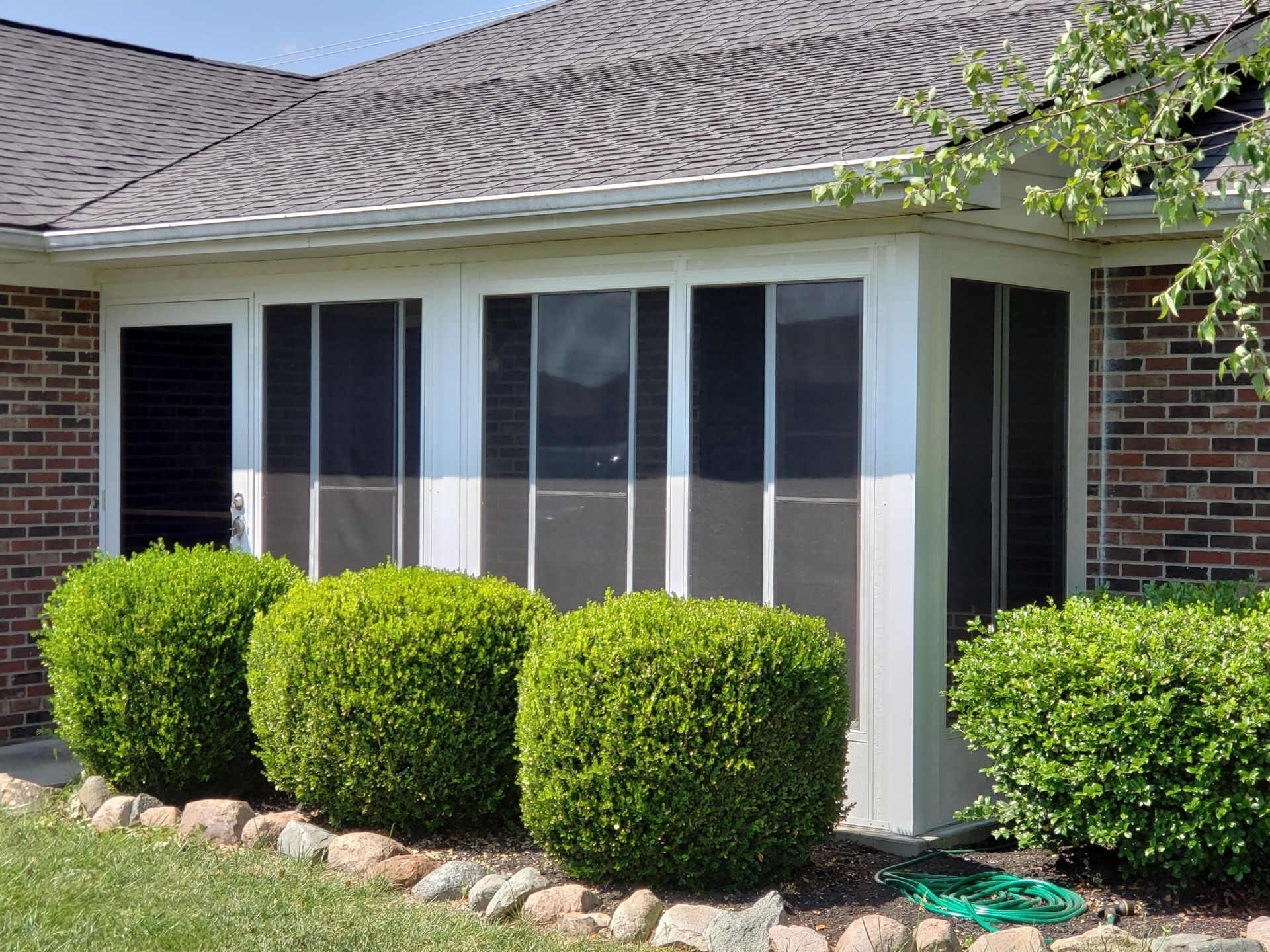 A sunroom addition with white frames and large screens attached to a brick house with round bushes in the foreground.
